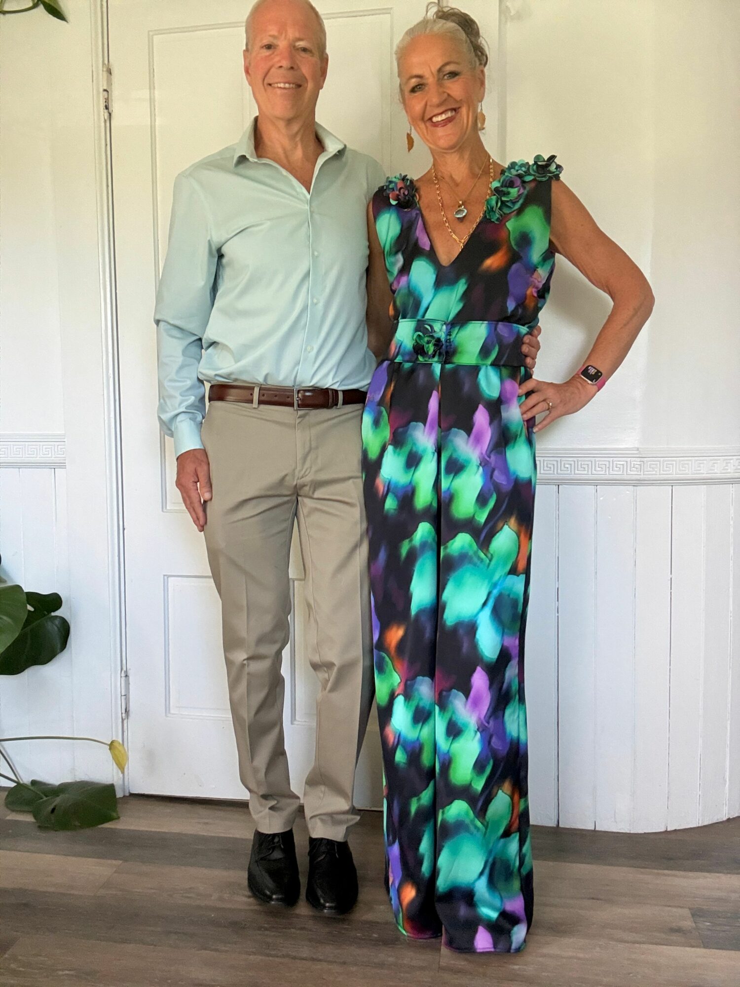 Two people standing indoors, one in a colorful dress and the other in a light shirt and beige pants, with a white wall background.