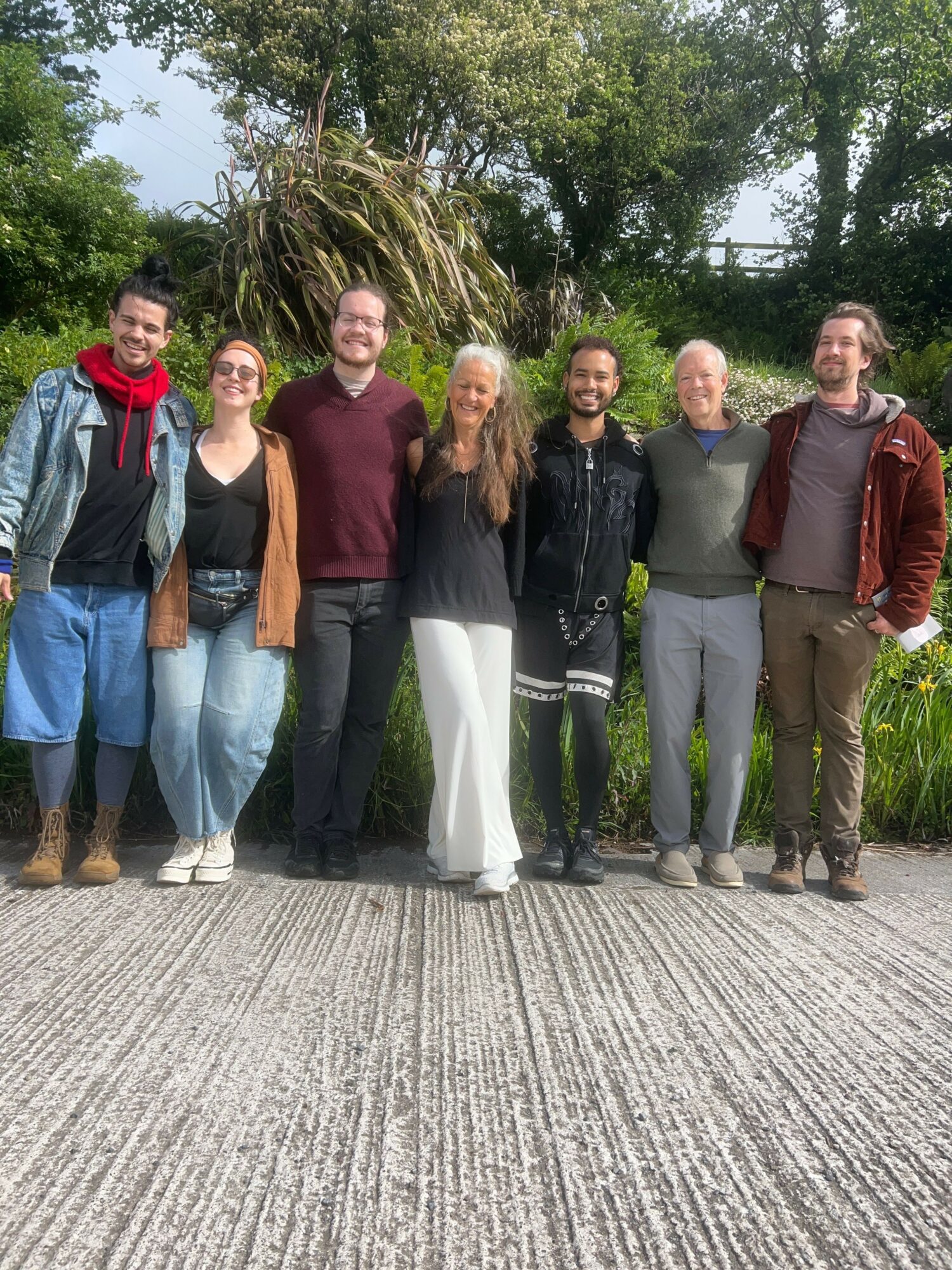 Six people standing outdoors on a paved path with trees and greenery in the background, smiling and posing for a photo.