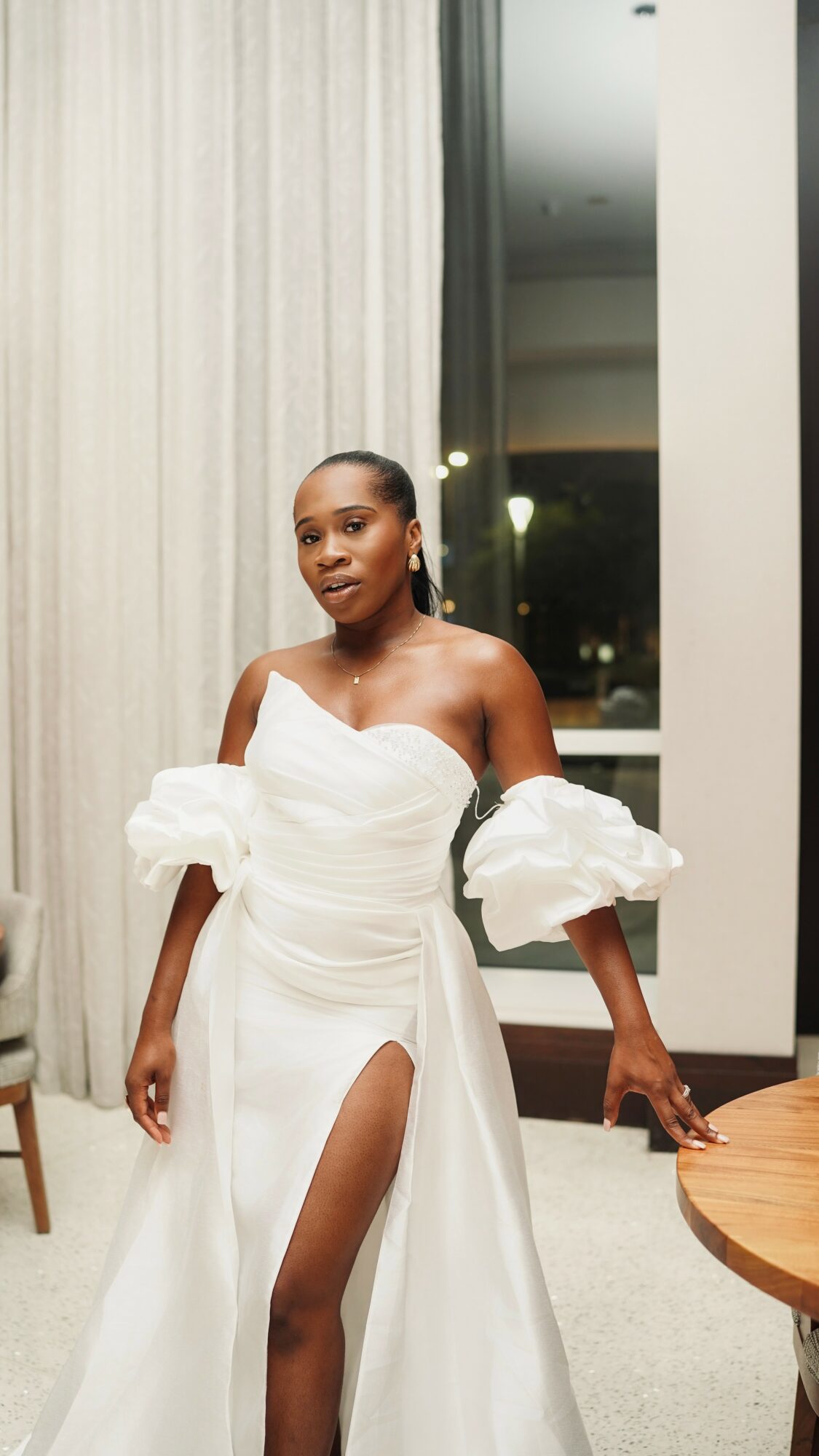Woman in white dress with off-shoulder sleeves and high slit, standing indoors near a window and curtain.