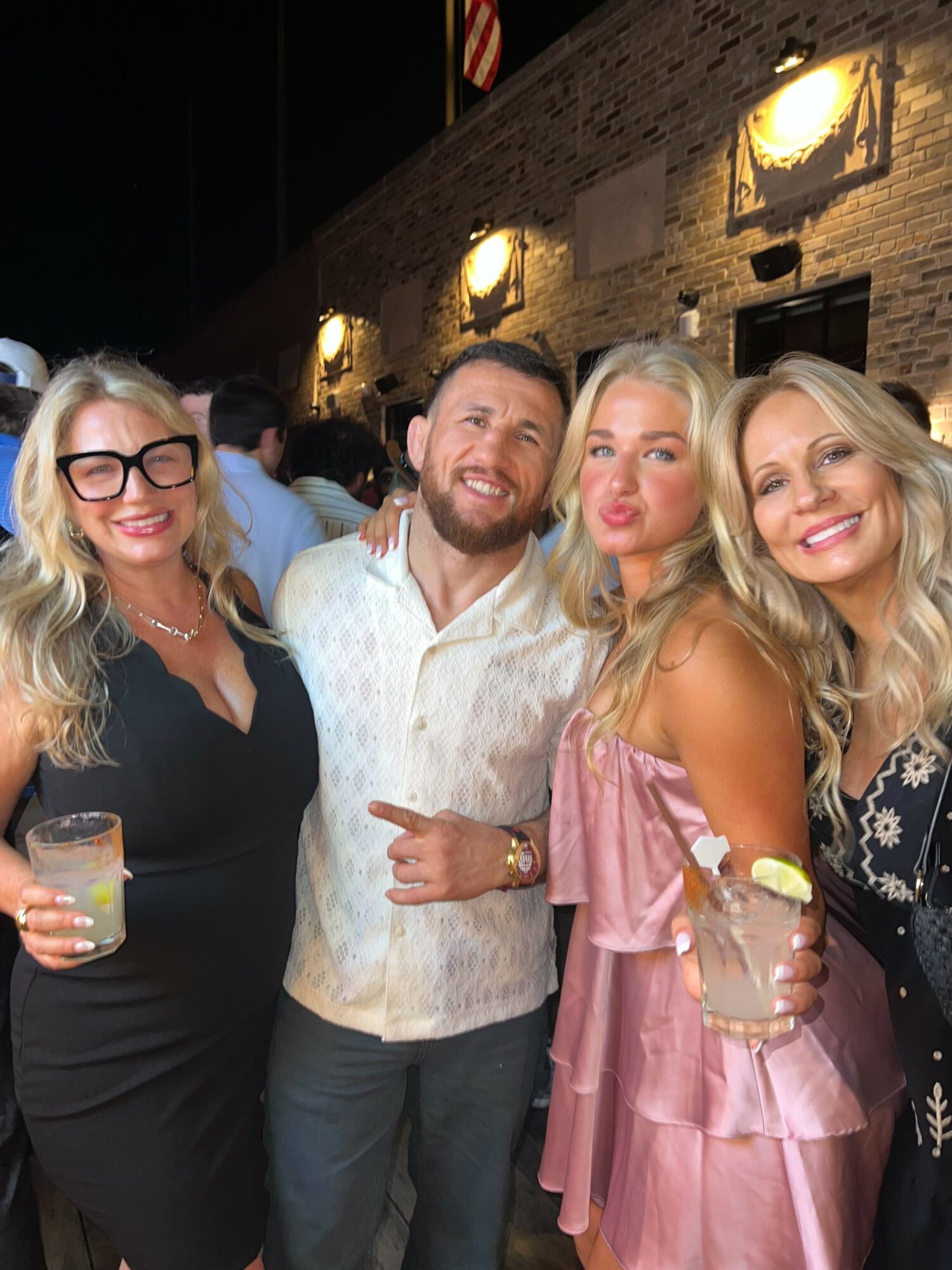 Four people smiling and posing together at a social event, holding drinks, in front of a brick wall with lights.