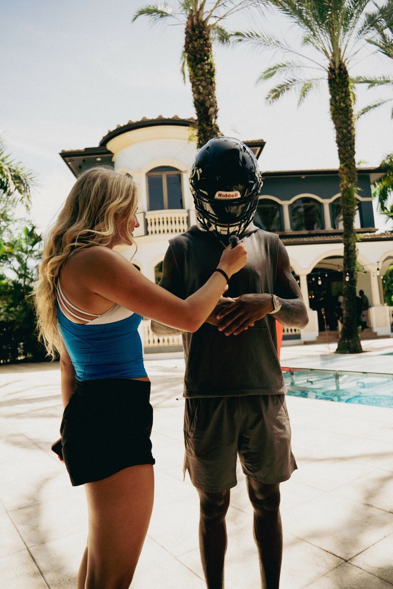 A woman helps a man with a motorcycle helmet outdoors near a pool, with palm trees and a building in the background.