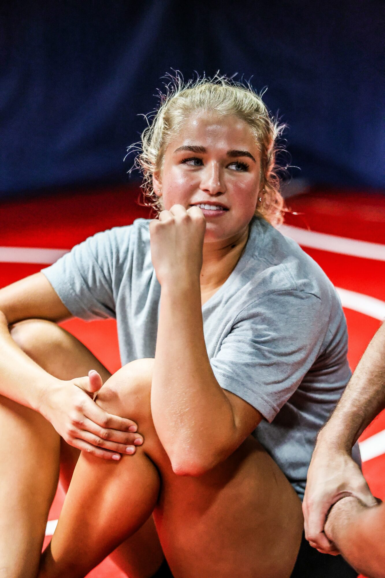 Young woman with blonde hair tied back, sitting on a red and white surface, resting her chin on her hand, smiling.