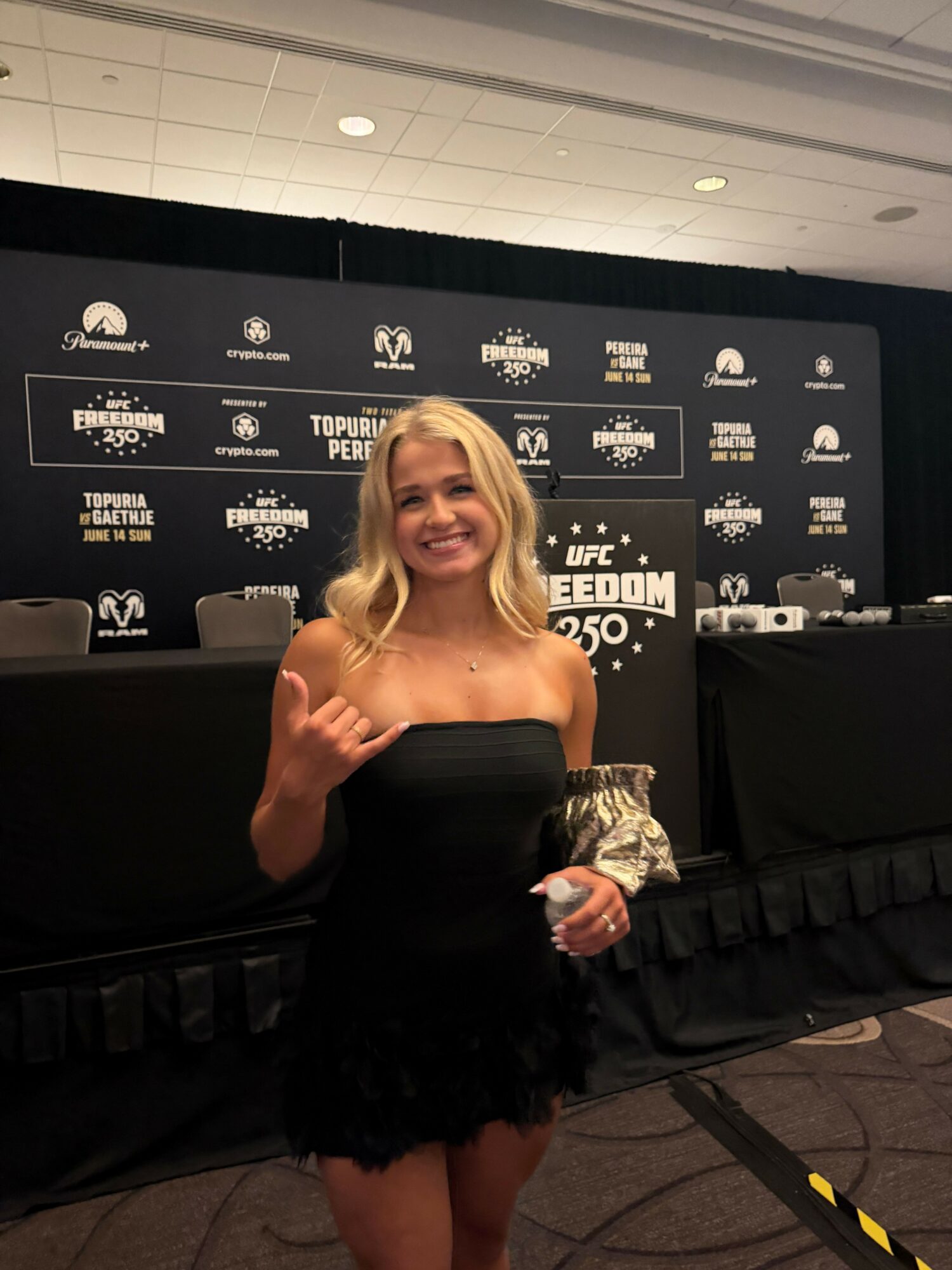 Woman in a black strapless dress smiling and giving a thumbs-up at an event, with a black table and backdrop behind her.