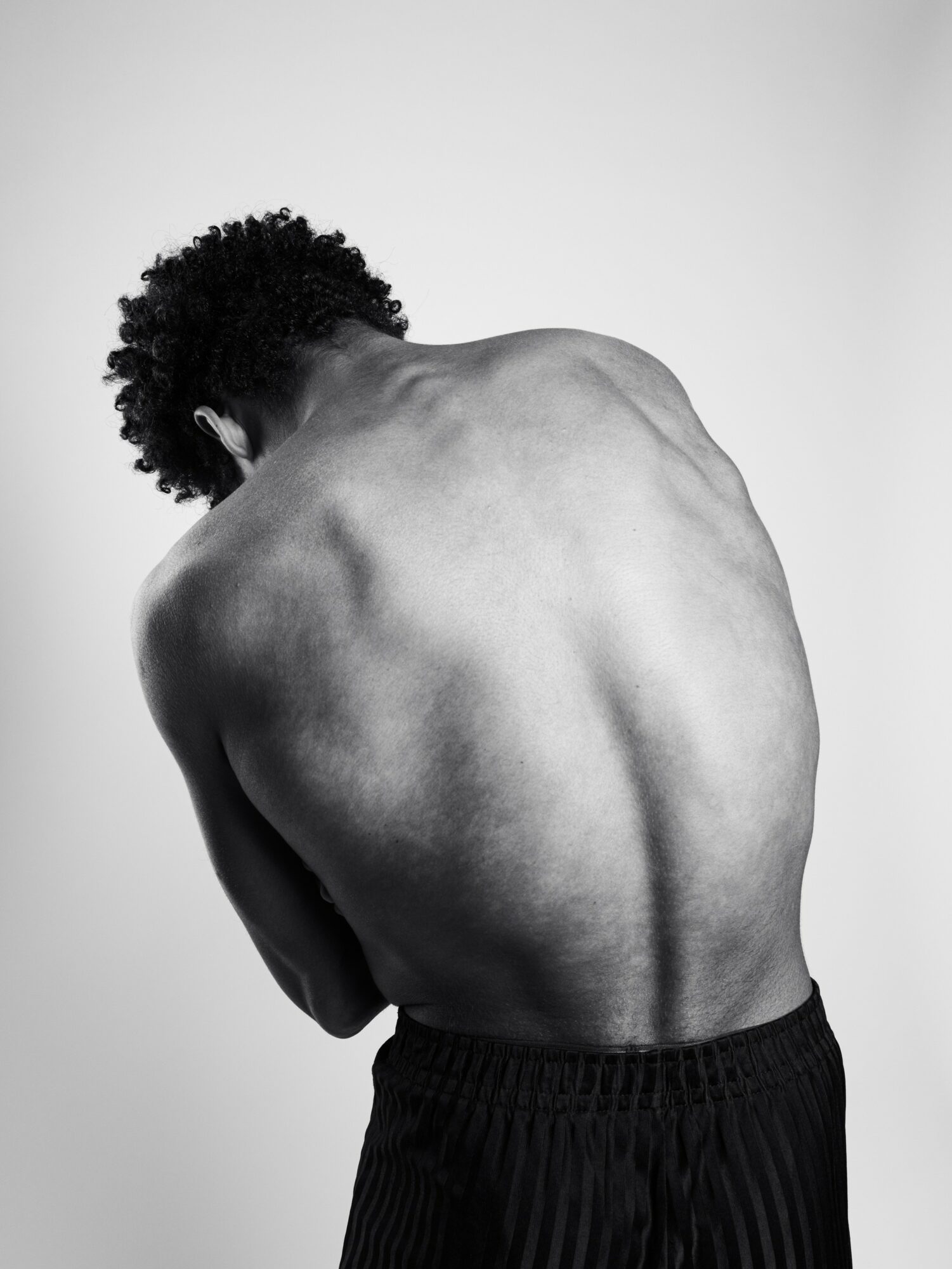 Back view of a person with curly hair, showing their bare back and shoulders, wearing dark pants, against a plain background.