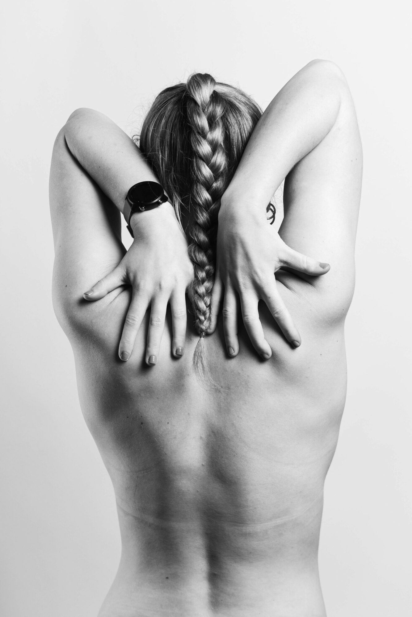Back view of a person with braided hair, arms raised, hands covering shoulders, in black and white.