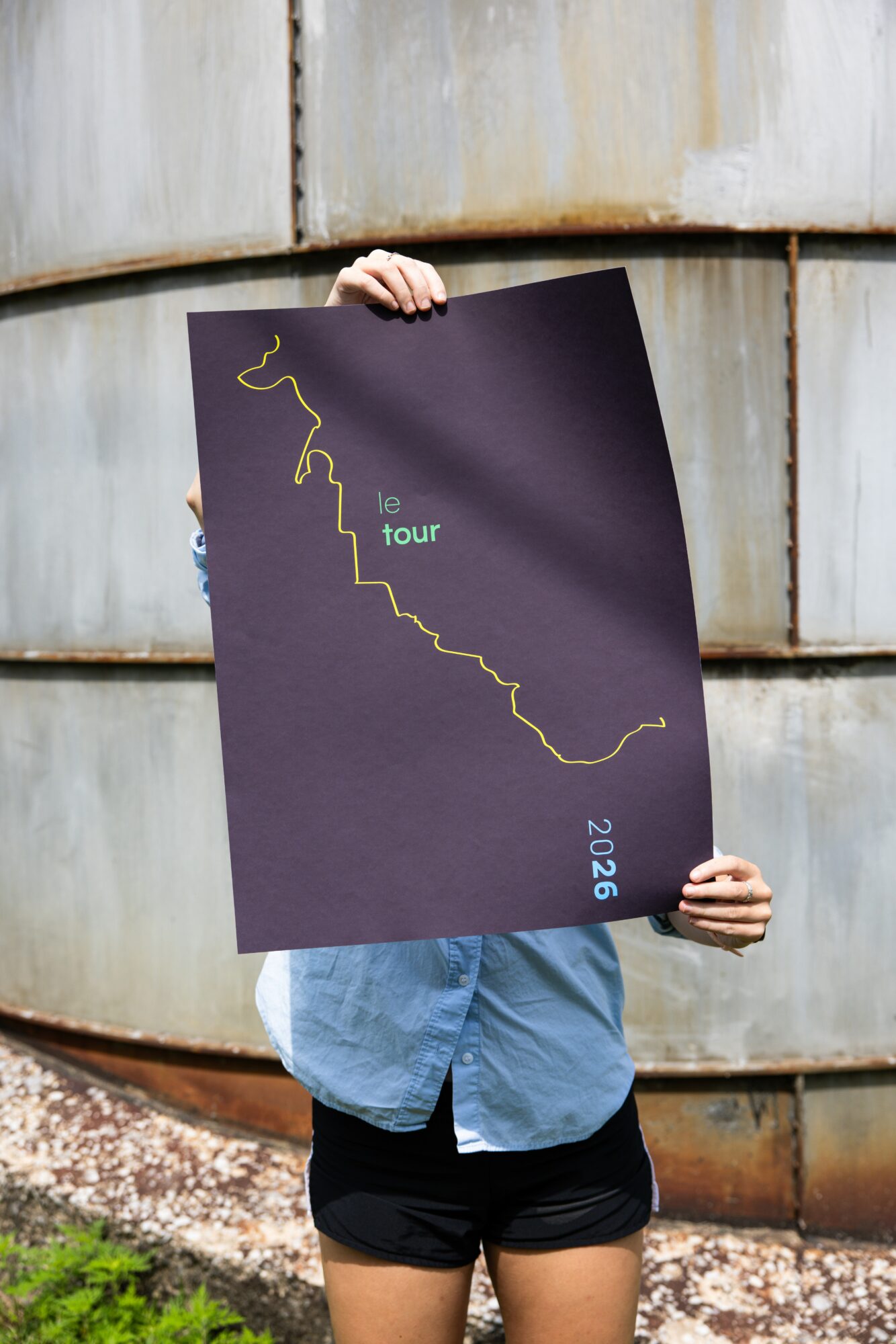 Person holding a dark poster with a yellow outline of a route and text, outdoors, rusted metal background, grass at bottom.