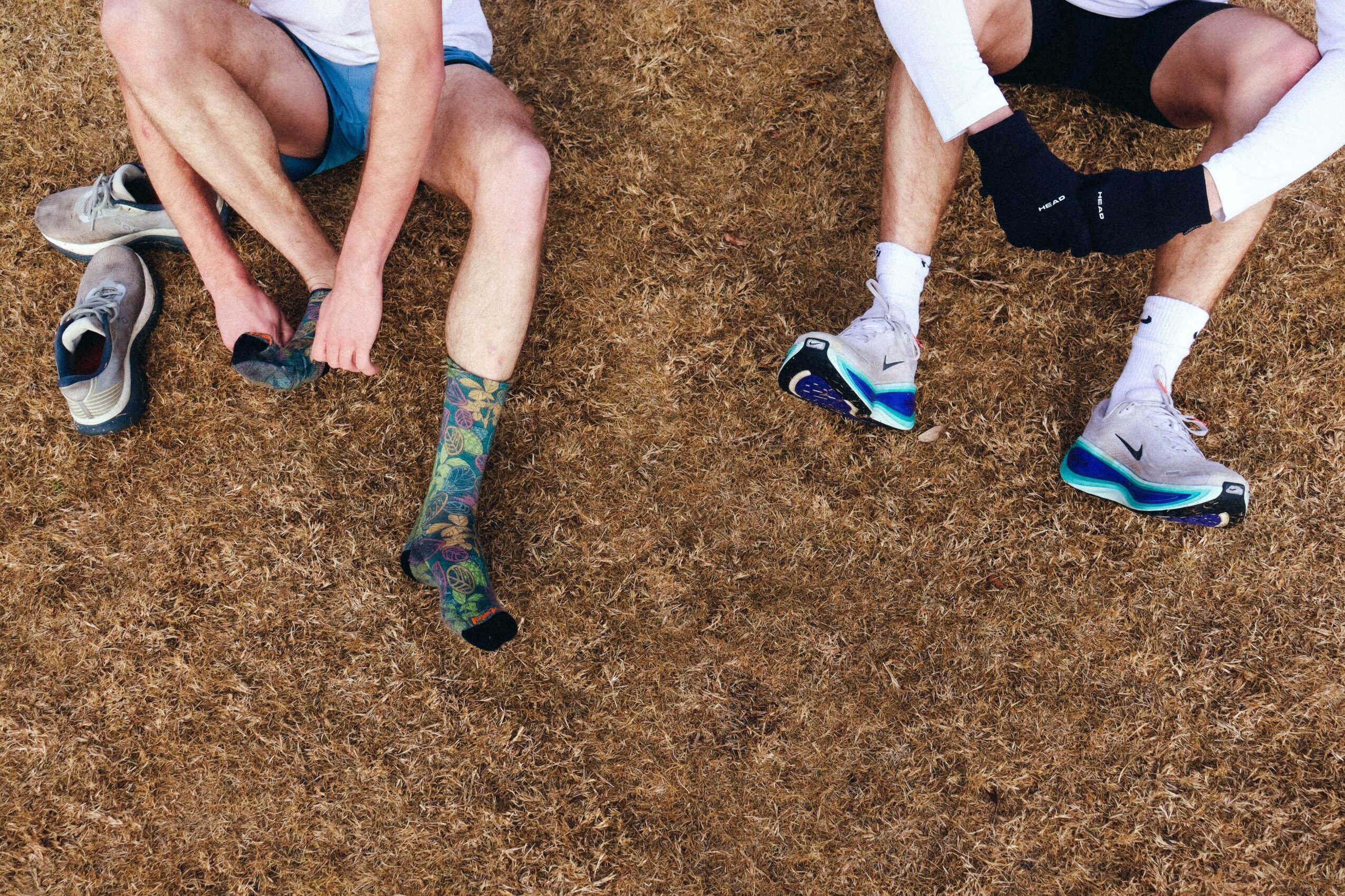 Two people sitting on the ground with legs extended, one wearing colorful socks and sneakers, the other in white socks and sneakers.