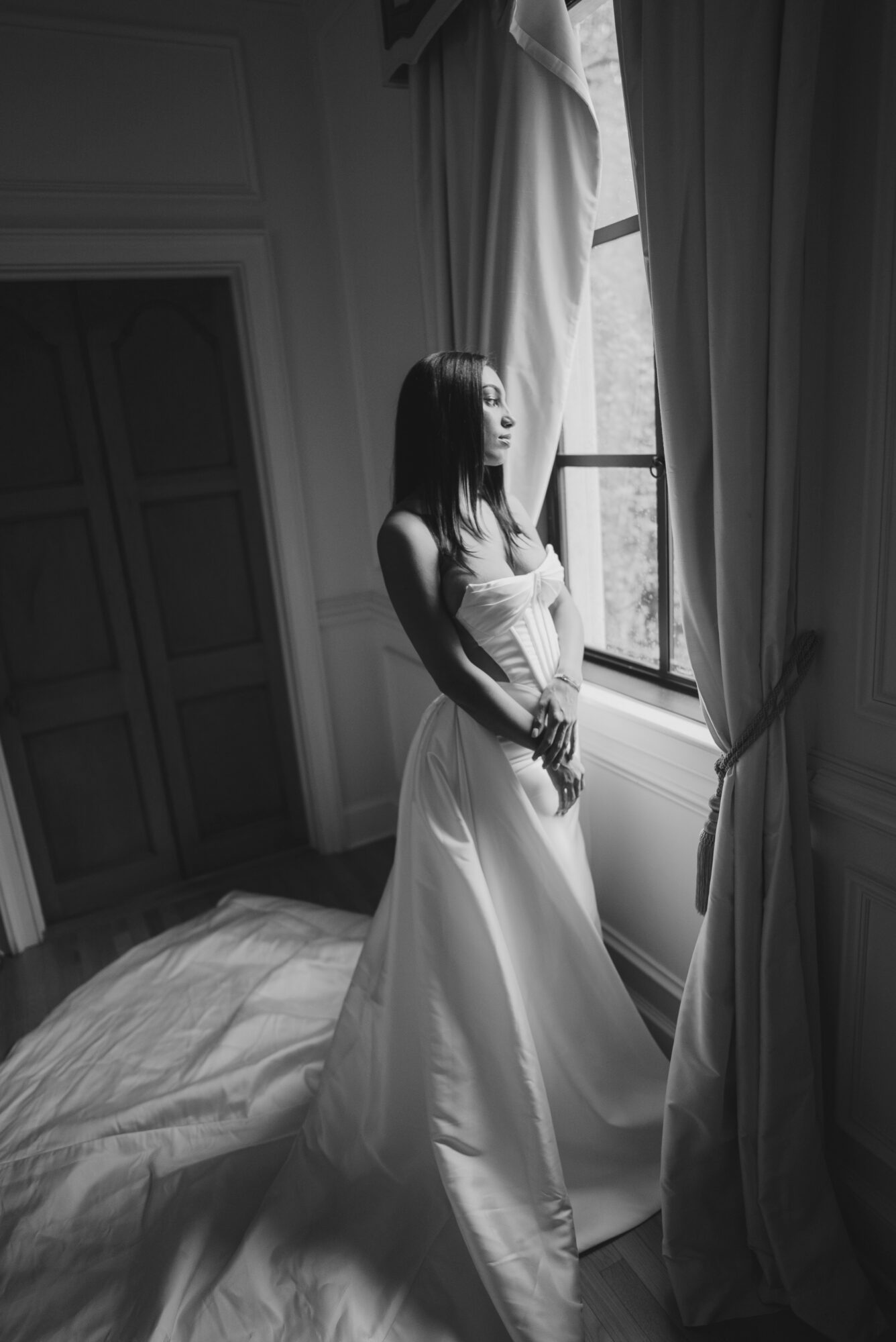 Woman in a long dress standing by a window with curtains, looking outside, in black and white.