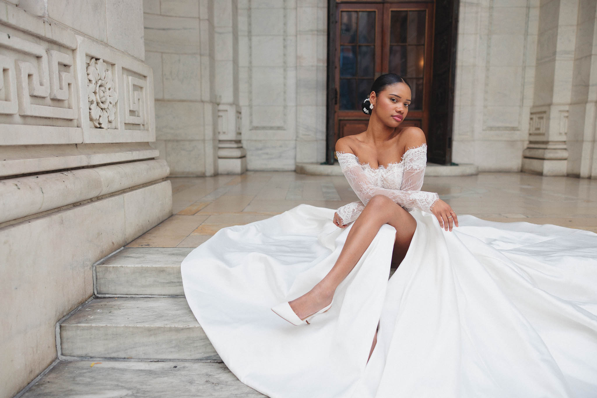 Woman in a white wedding dress sitting on marble steps in a grand building interior.