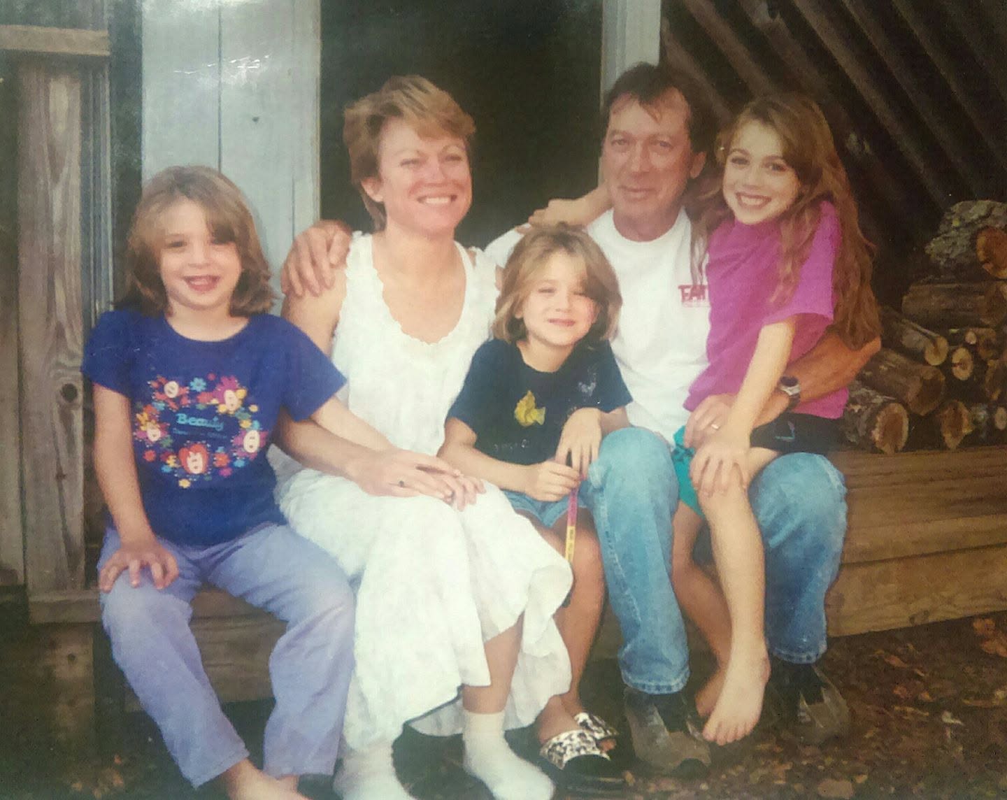 Family of six sitting together outdoors, smiling, with a wooden structure and logs in background.