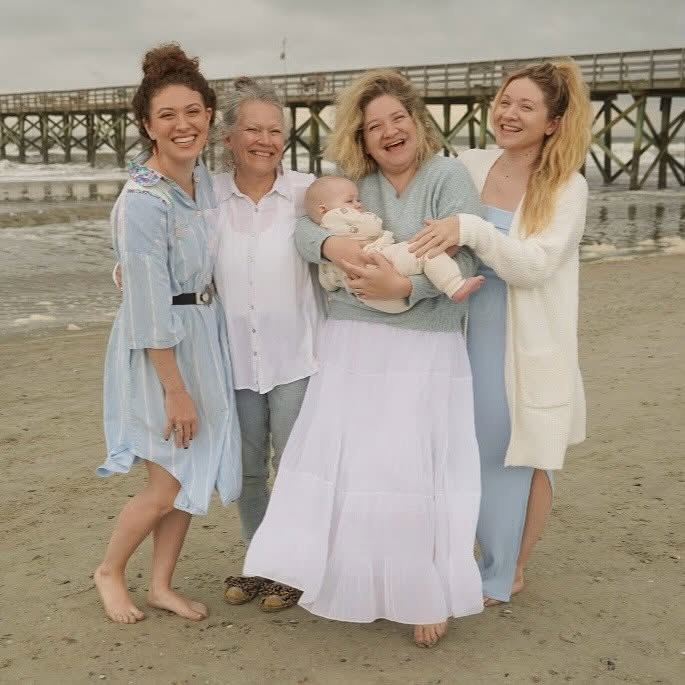 Five women and a baby standing on a beach near a pier, smiling and holding the baby, with ocean in background.