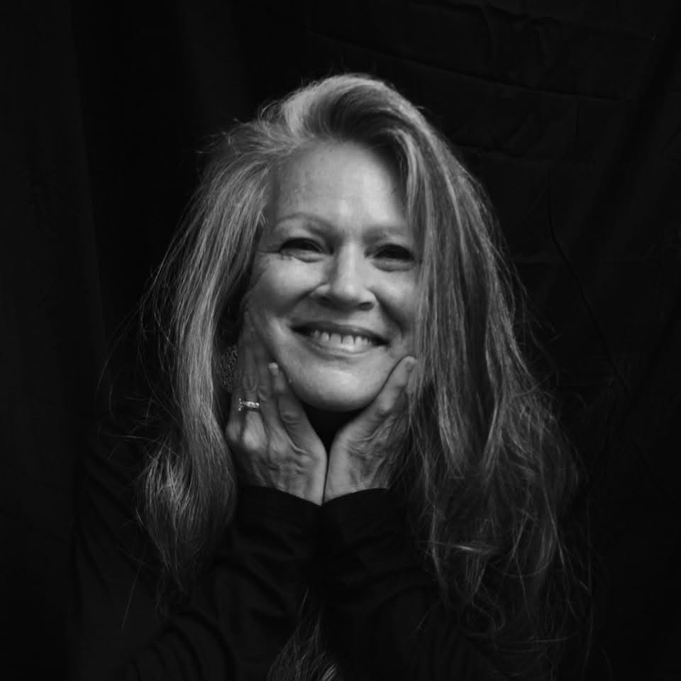Smiling woman with long hair, hands on face, black background, black clothing, cheerful expression.
