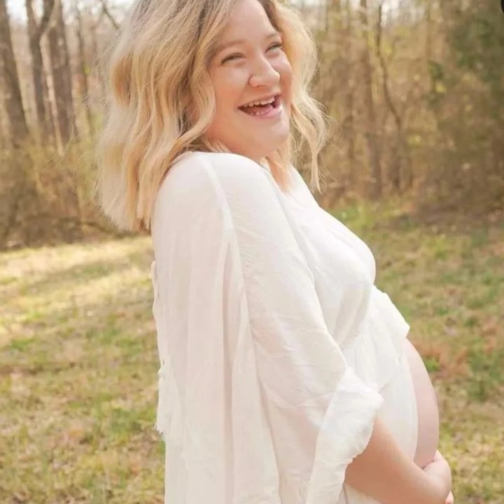 Smiling pregnant woman with blonde hair outdoors in a wooded area, wearing a white shirt, looking to the side.