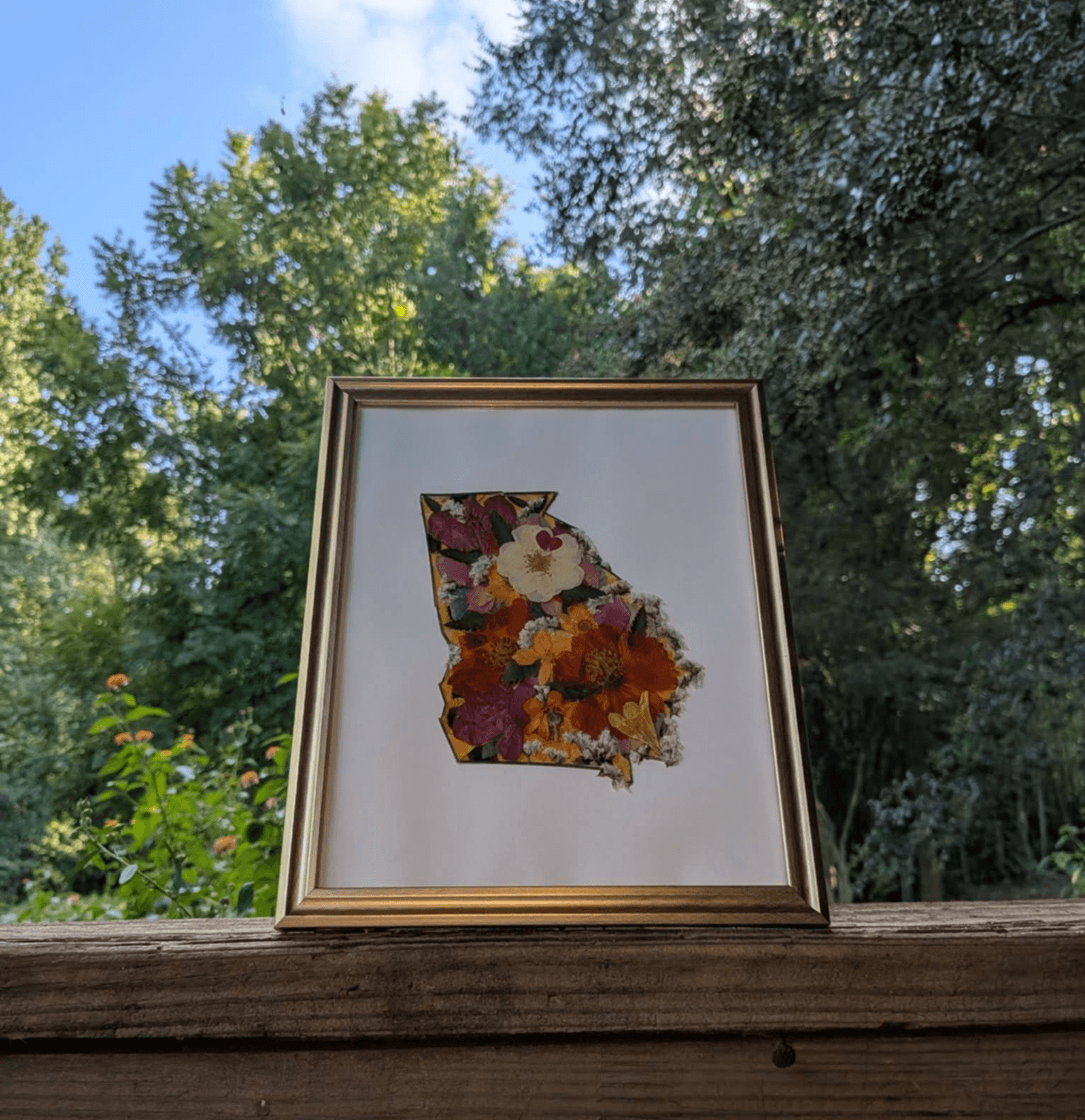 Framed artwork of the state of Georgia filled with colorful flowers, placed outdoors on a wooden surface with trees and sky in background.