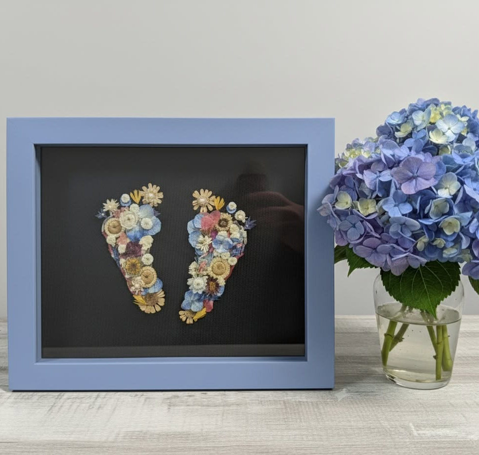 Framed artwork of two feet made from colorful flowers, next to a vase of blue hydrangeas on a white surface.