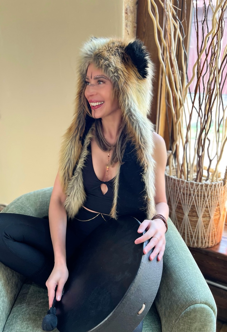 Woman smiling, wearing a furry animal hat, sitting on a green couch, with a large decorative plant nearby.