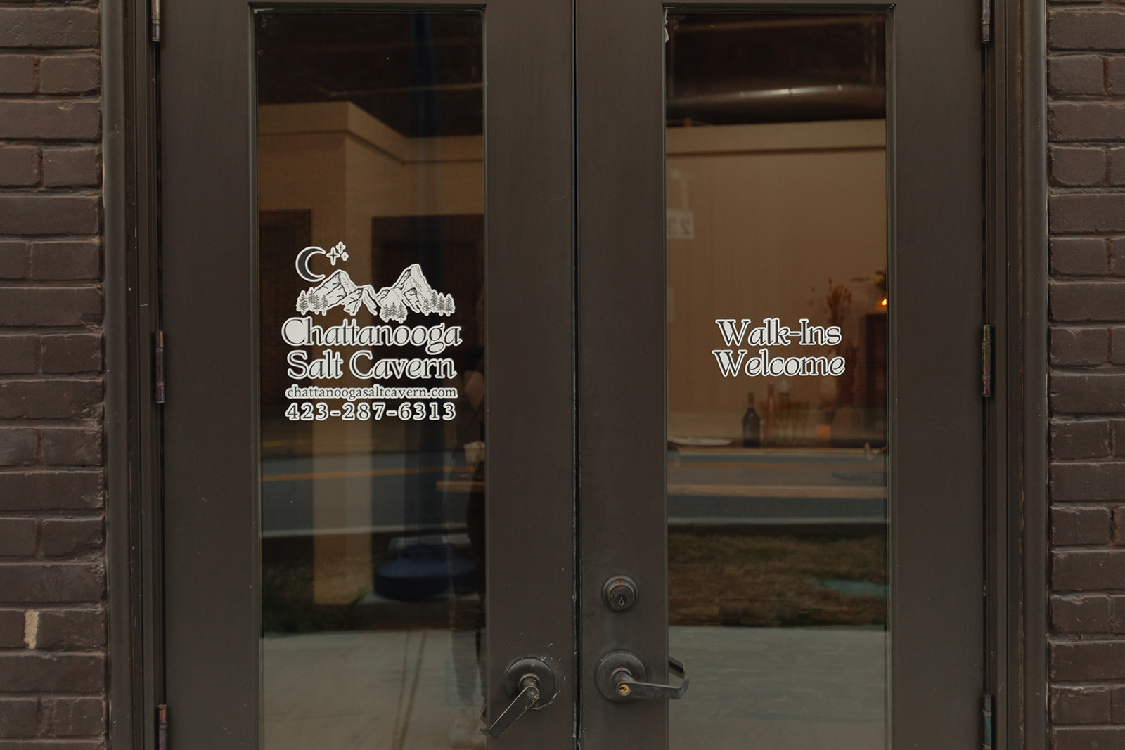 Glass door with white text and logo, brick wall on sides, reflections visible on glass, door handles in center.