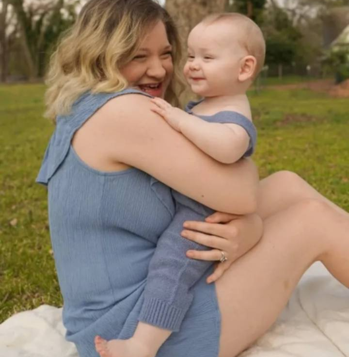 Woman holding smiling baby outdoors on grass, both wearing blue clothing, woman with blonde hair, baby with light hair.