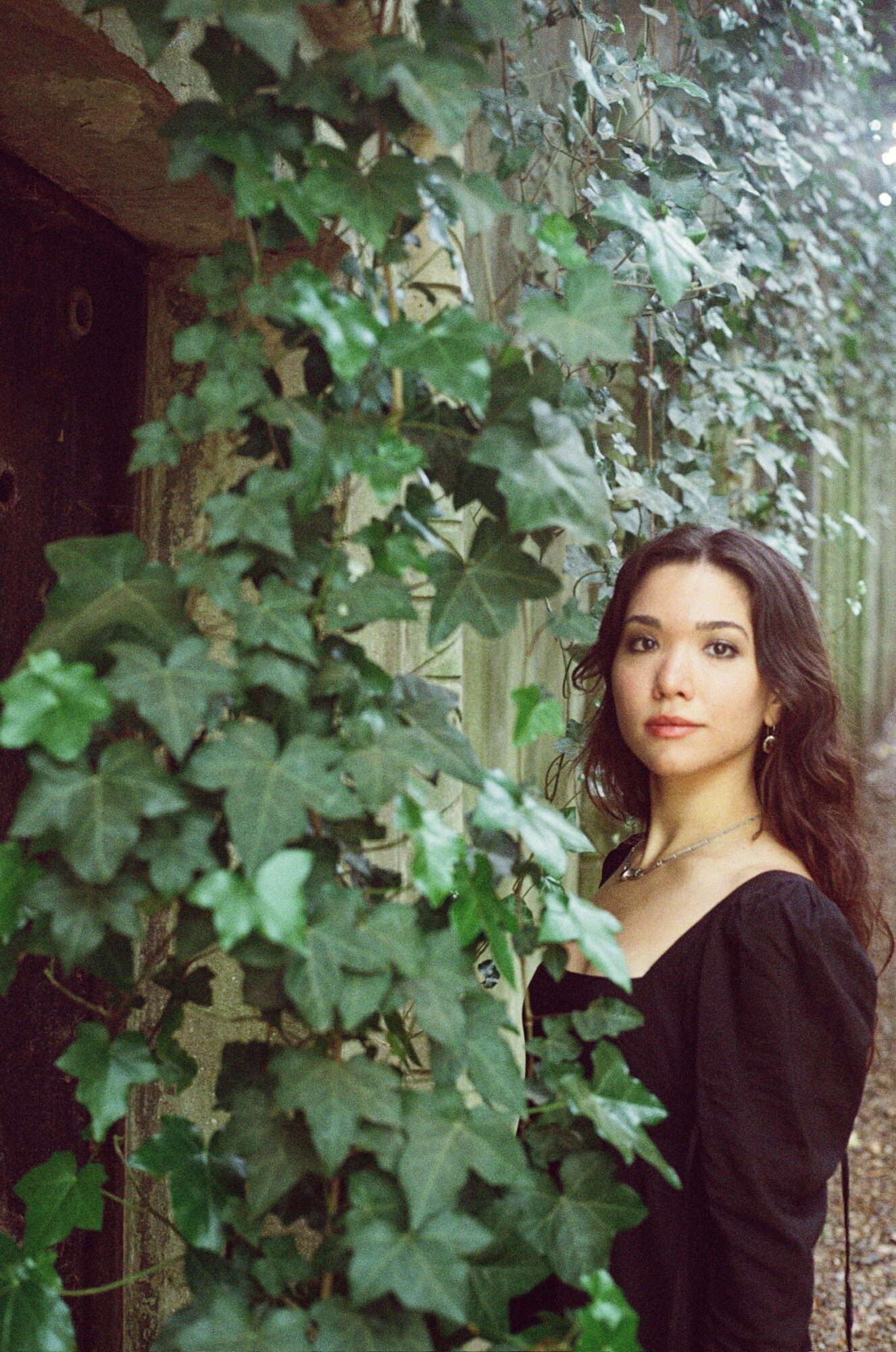 Young woman with dark wavy hair and light makeup standing among green ivy leaves outdoors.