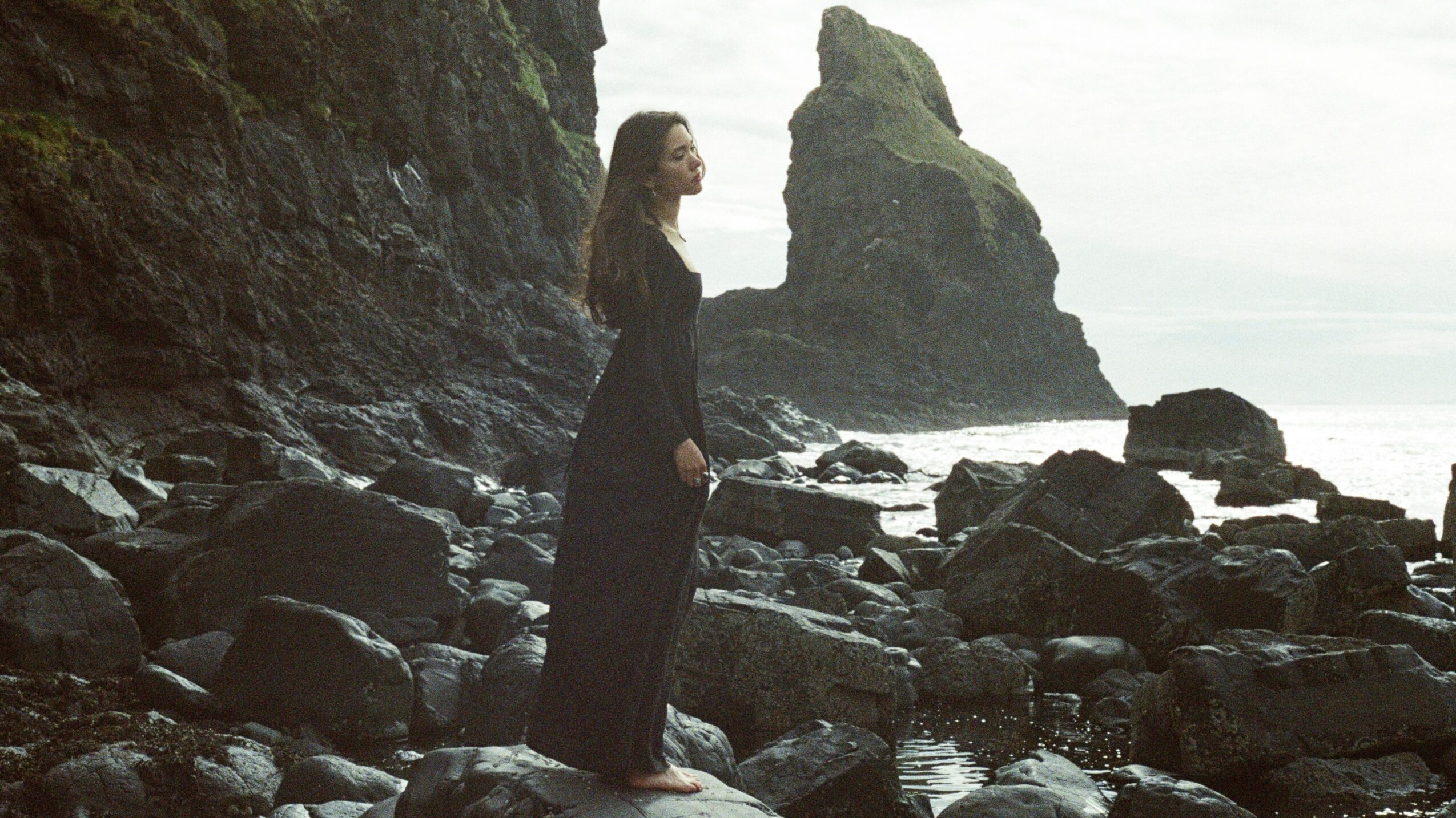 Woman standing on rocky shoreline with cliffs and sea in background, facing left, wearing dark clothing.