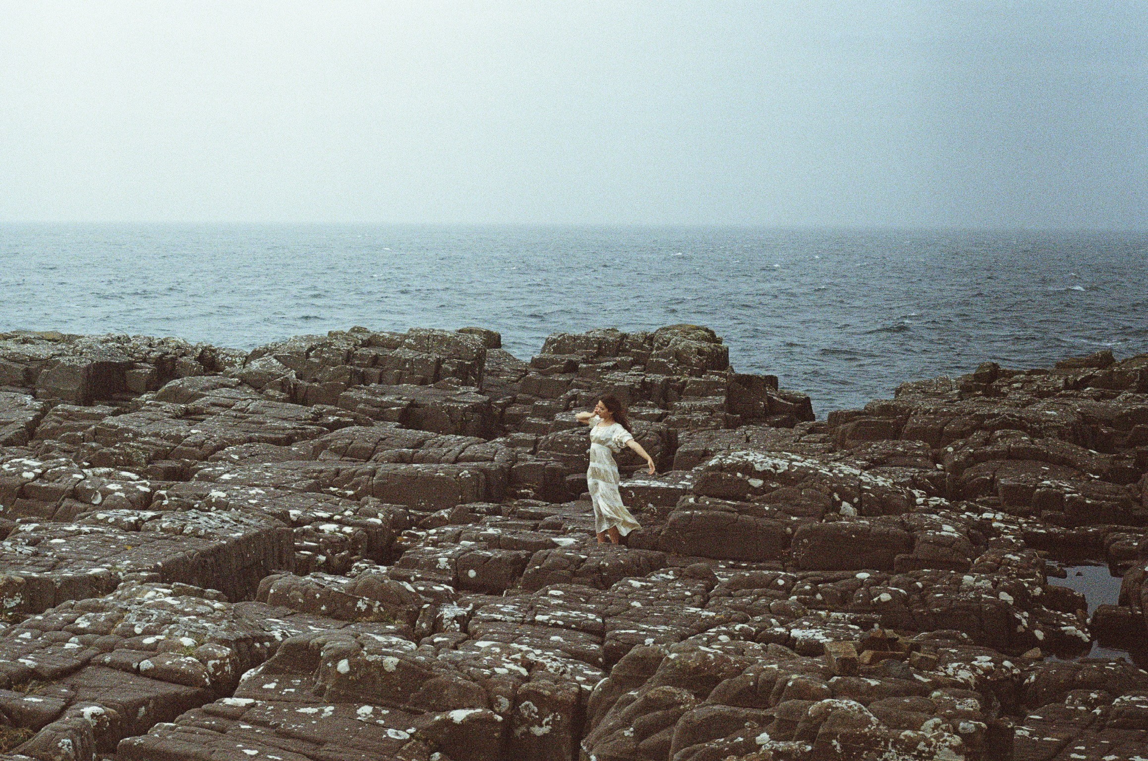Person standing on rocky shoreline near the ocean, facing the water, with cloudy sky above.