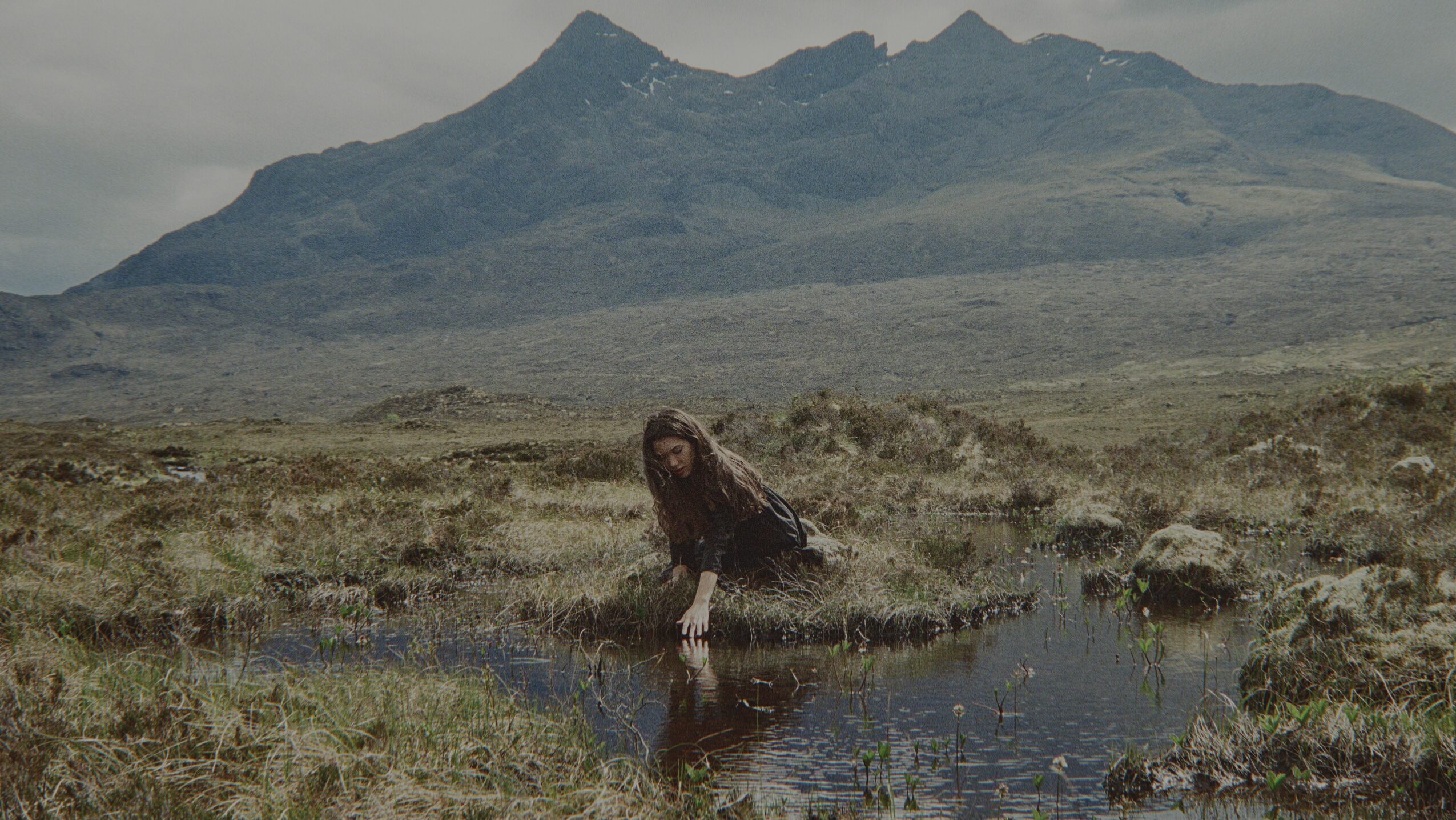 Person crouching near a small water body in a grassy landscape with mountains in the background.