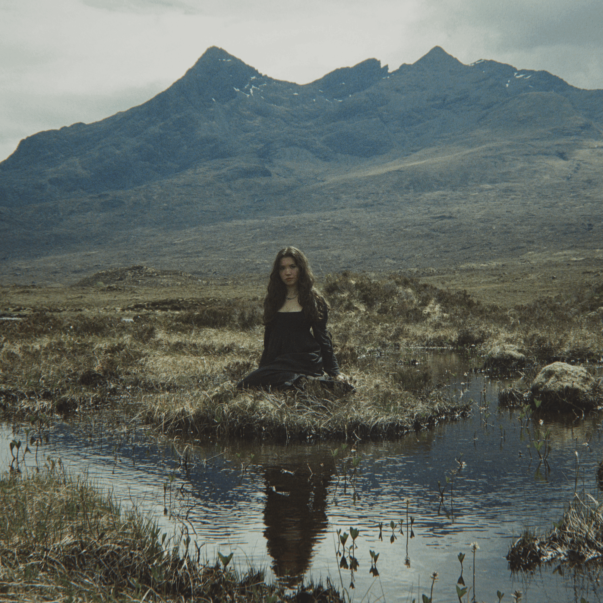 A woman with long hair sits on a small grassy island in a pond, mountains in the background, cloudy sky.
