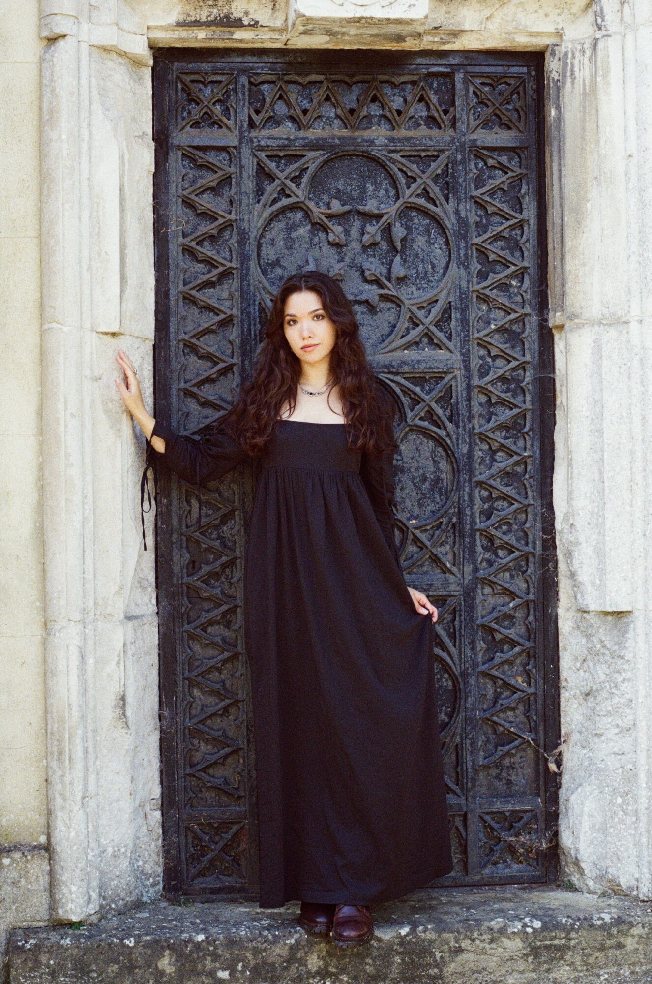 Woman in a long black dress standing in front of an ornate black metal gate, holding the side of the gate.