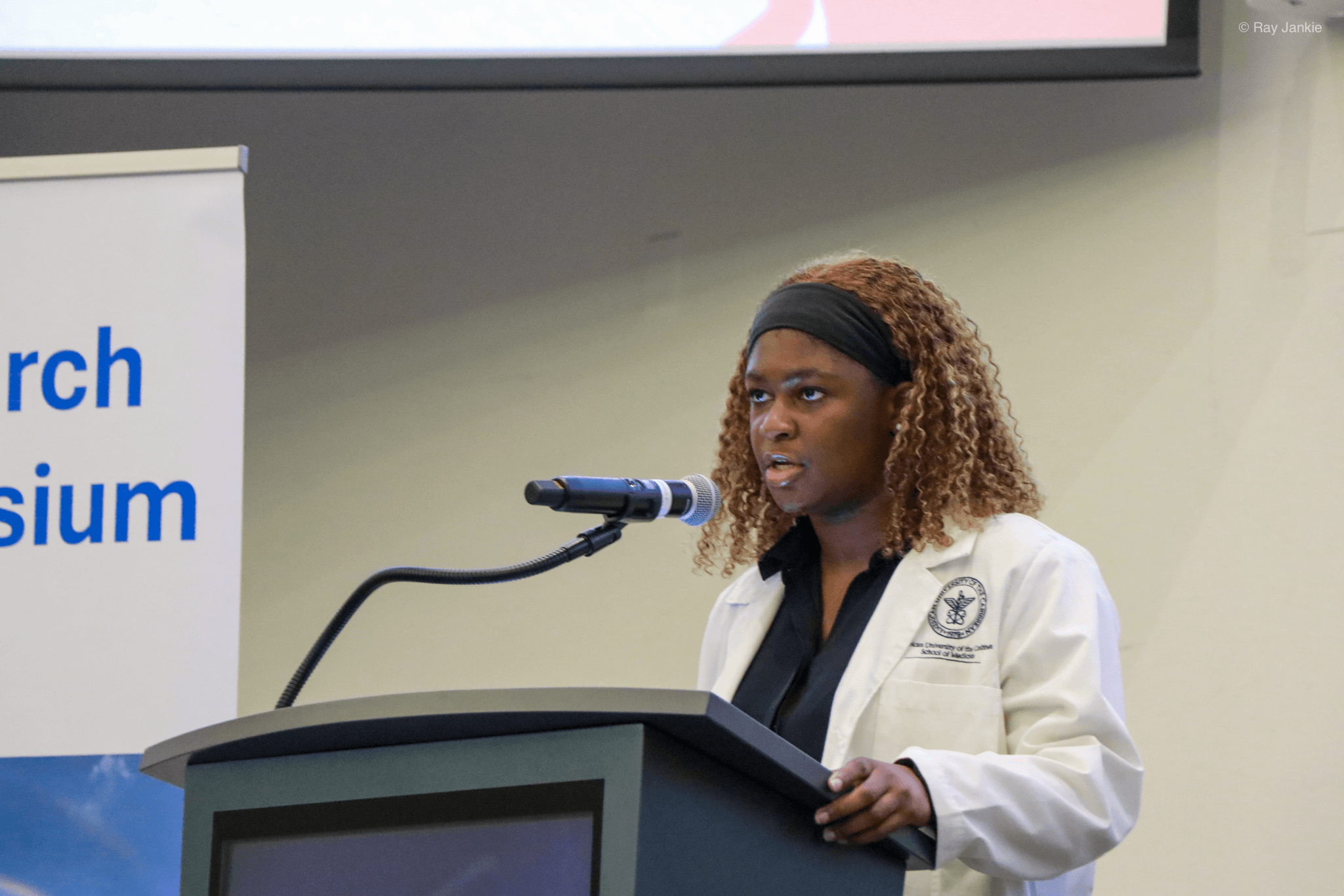 Woman speaking at a podium with a microphone, wearing a white coat, in a conference room setting.