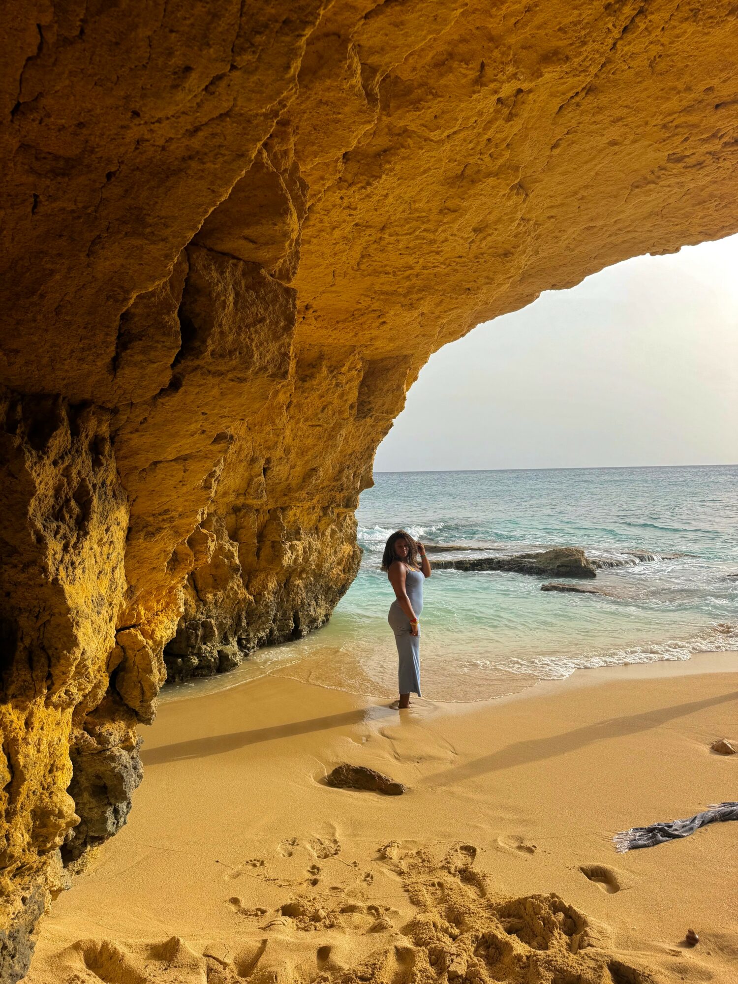 Person standing on sandy beach near ocean, under large rock formation, with sky and water in background.