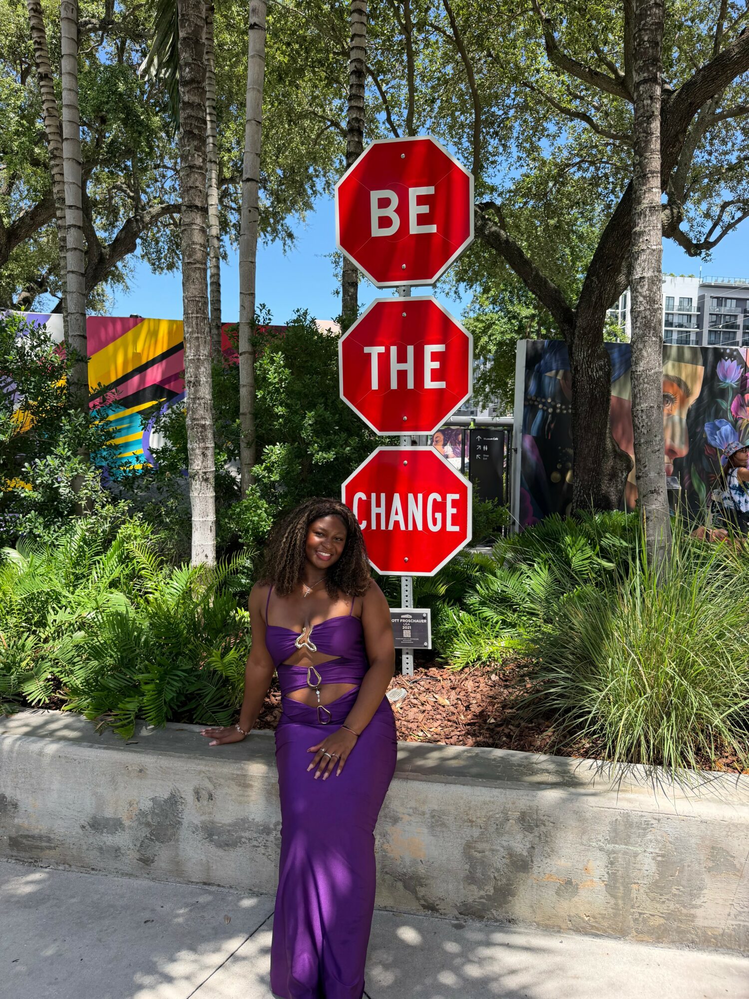 Woman in purple dress standing in front of a sign with three red stop signs reading 'BE THE CHANGE'.