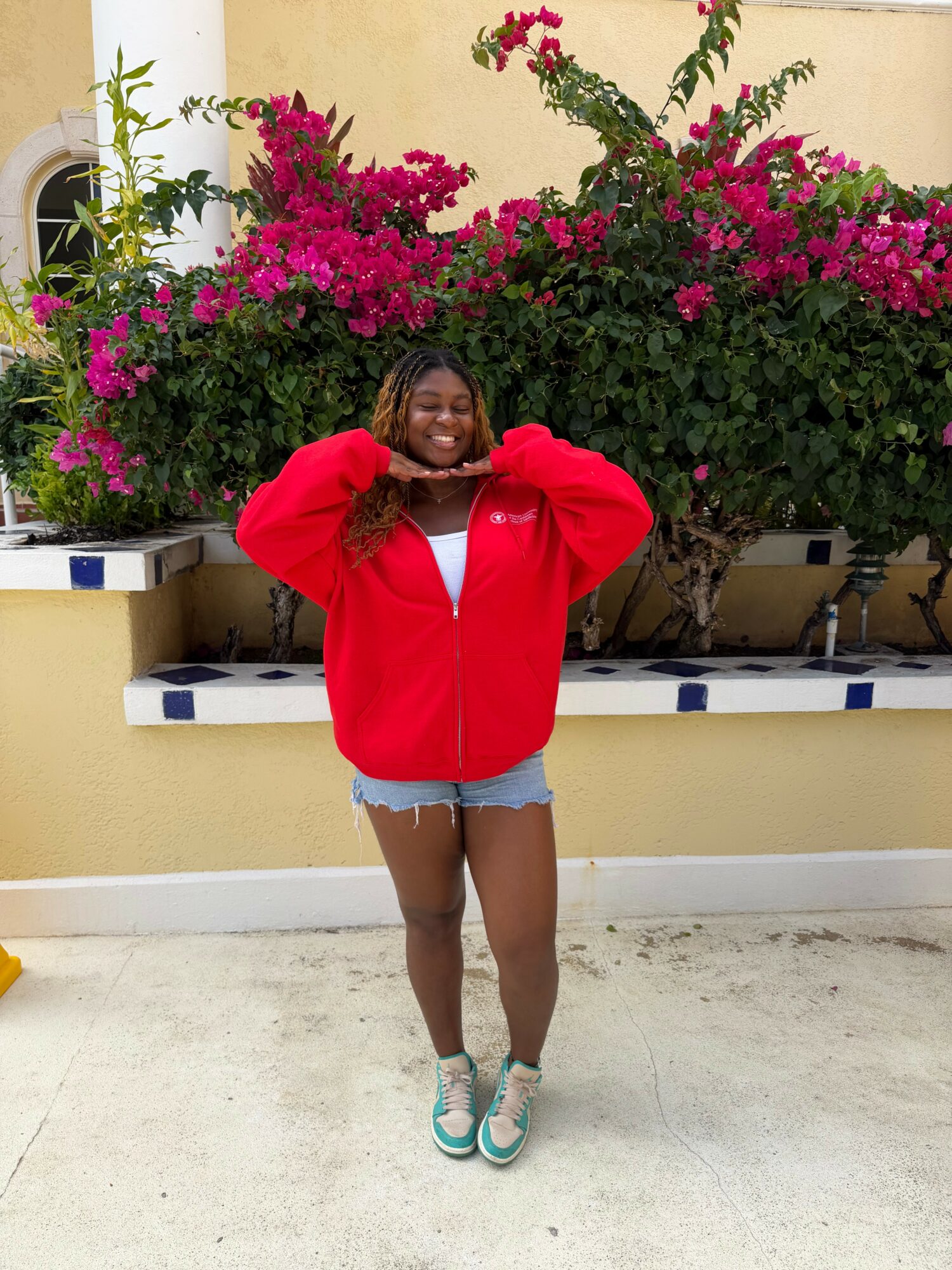Person standing outdoors in front of a flowering bush, smiling, wearing a red jacket, denim shorts, and sneakers.