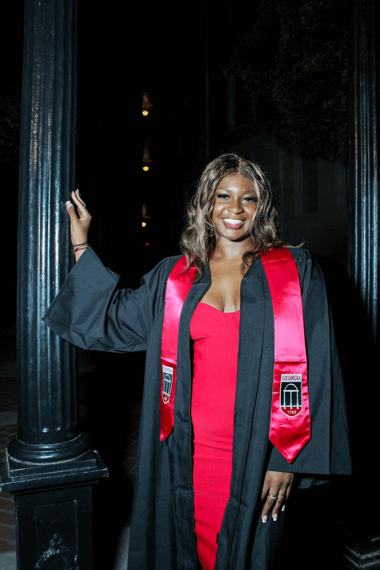 Woman in graduation gown and red dress smiling, standing next to a column, holding onto it, in a dark setting.