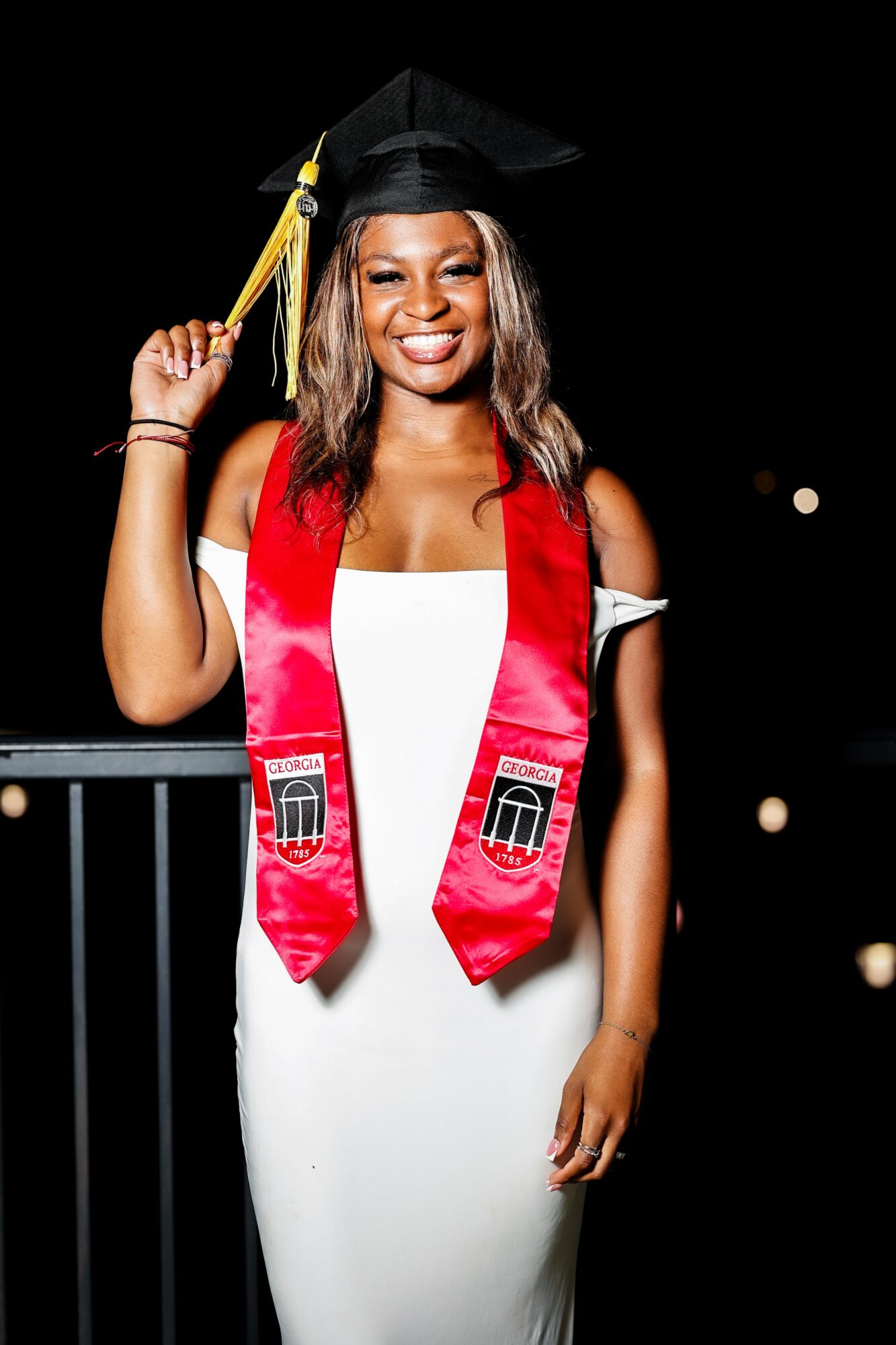 Woman in graduation cap and gown with red stole, smiling, holding her cap, night background with lights, 20 words.