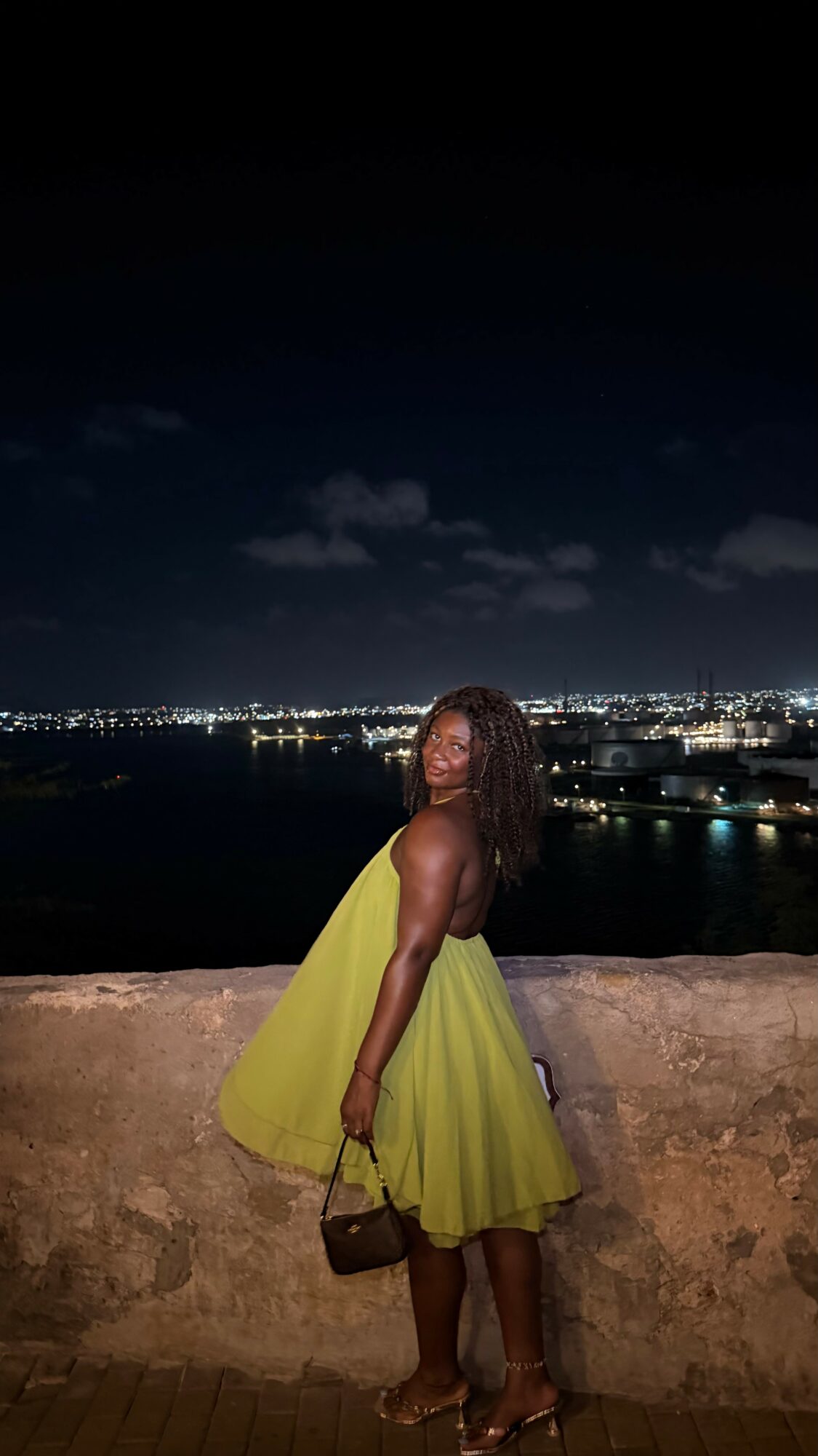 Woman in yellow dress standing by a stone wall at night with city lights in the background.