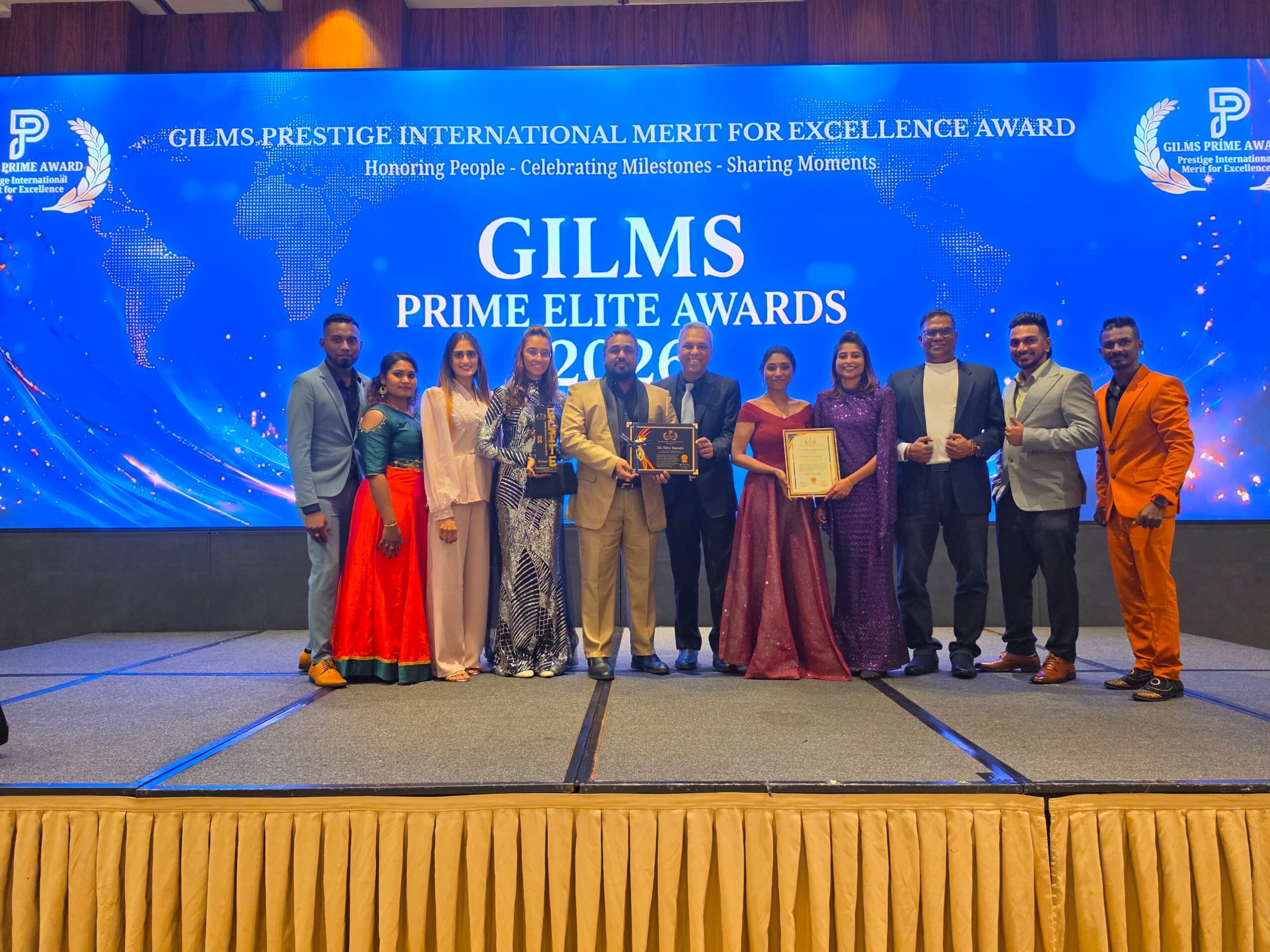 Group of people on stage holding awards at Gilms Prime Elite Awards ceremony, blue background with event details, yellow curtain at bottom.