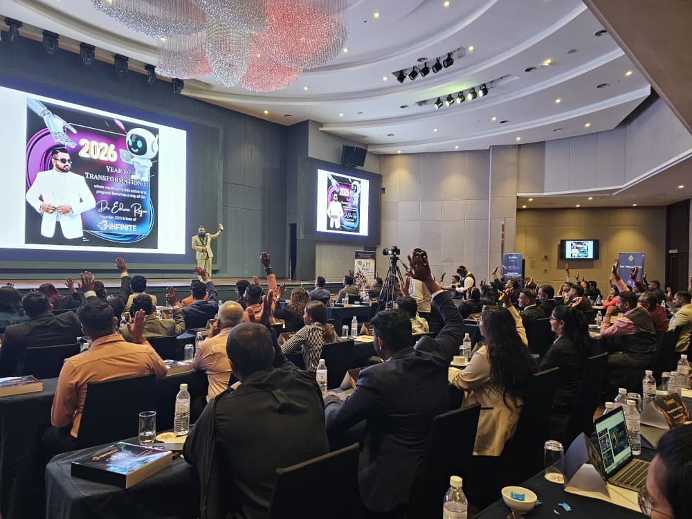 Conference room filled with seated audience watching a presentation on large screens, with a speaker on stage and decorative lighting.