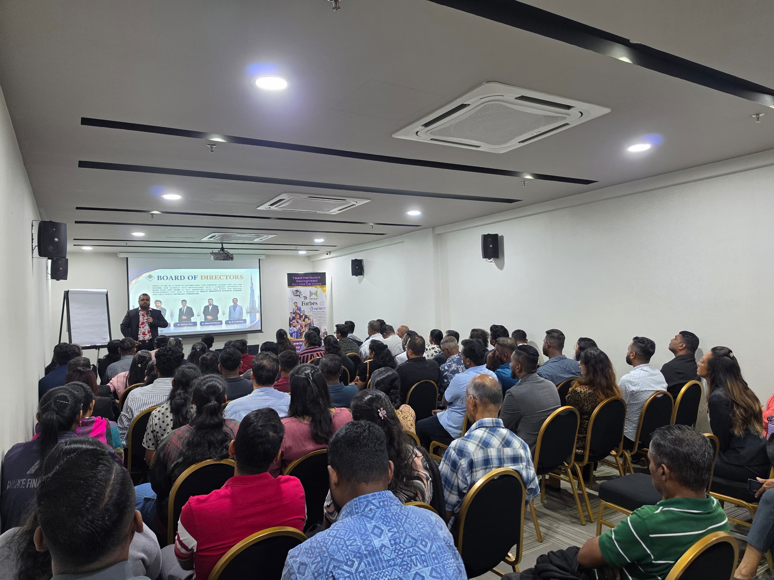 Audience seated in a conference room watching a presentation on a screen at the front.