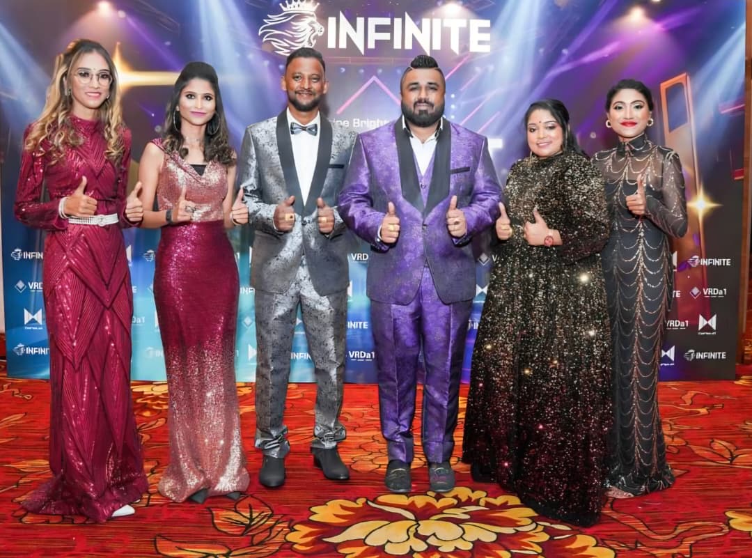 Group of seven people in formal attire standing on a stage with a backdrop reading 'INFINITE'.
