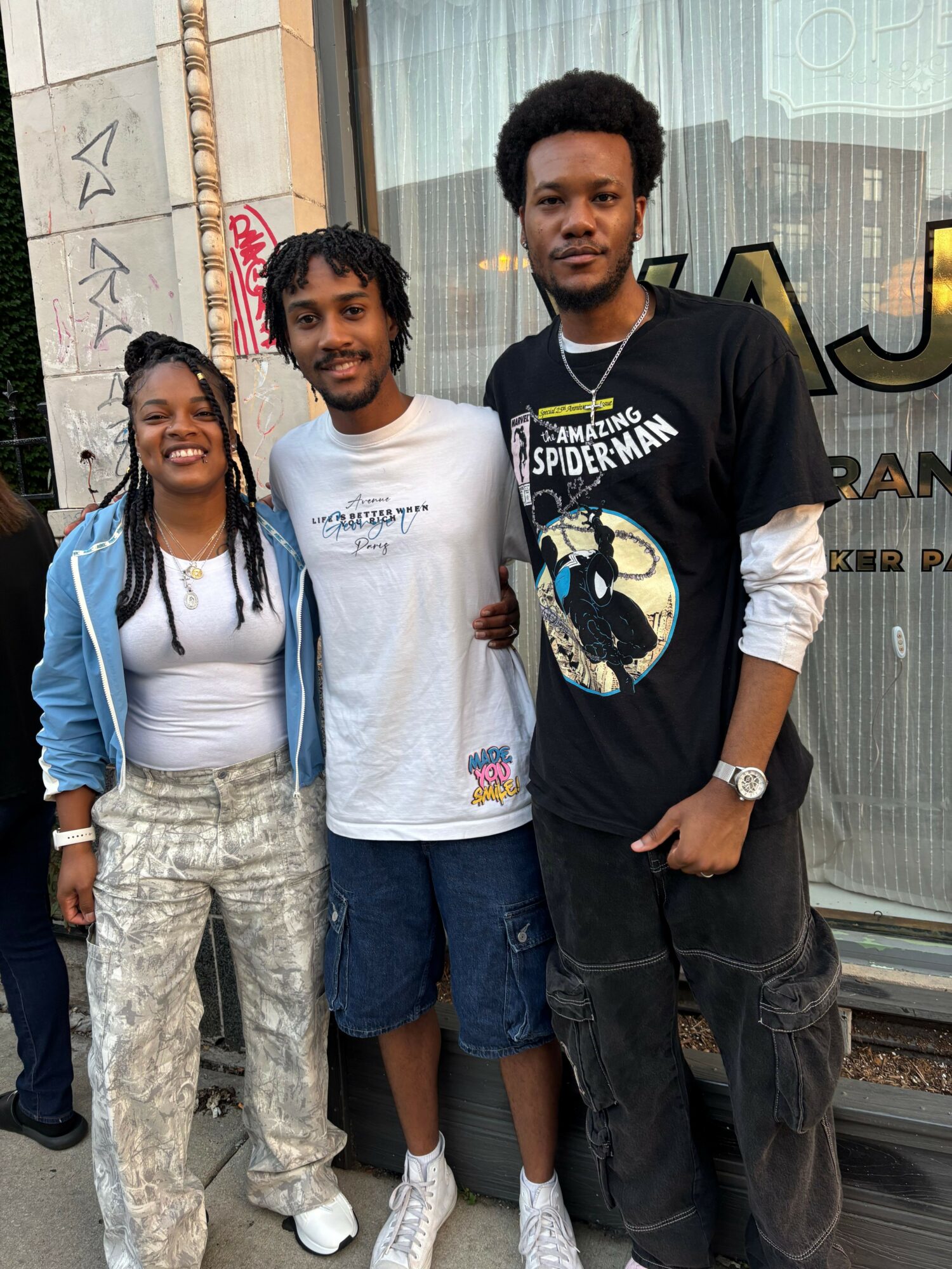 Three people standing outdoors in front of a building, two men and one woman, smiling and posing for the photo.