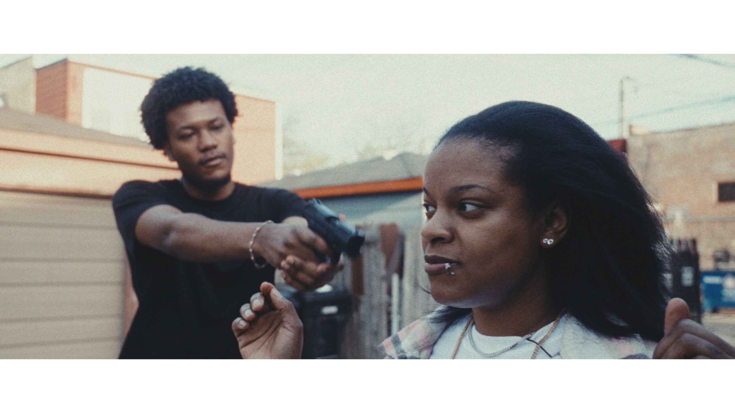 A young man points a gun at a woman with long dark hair outdoors in an urban setting.