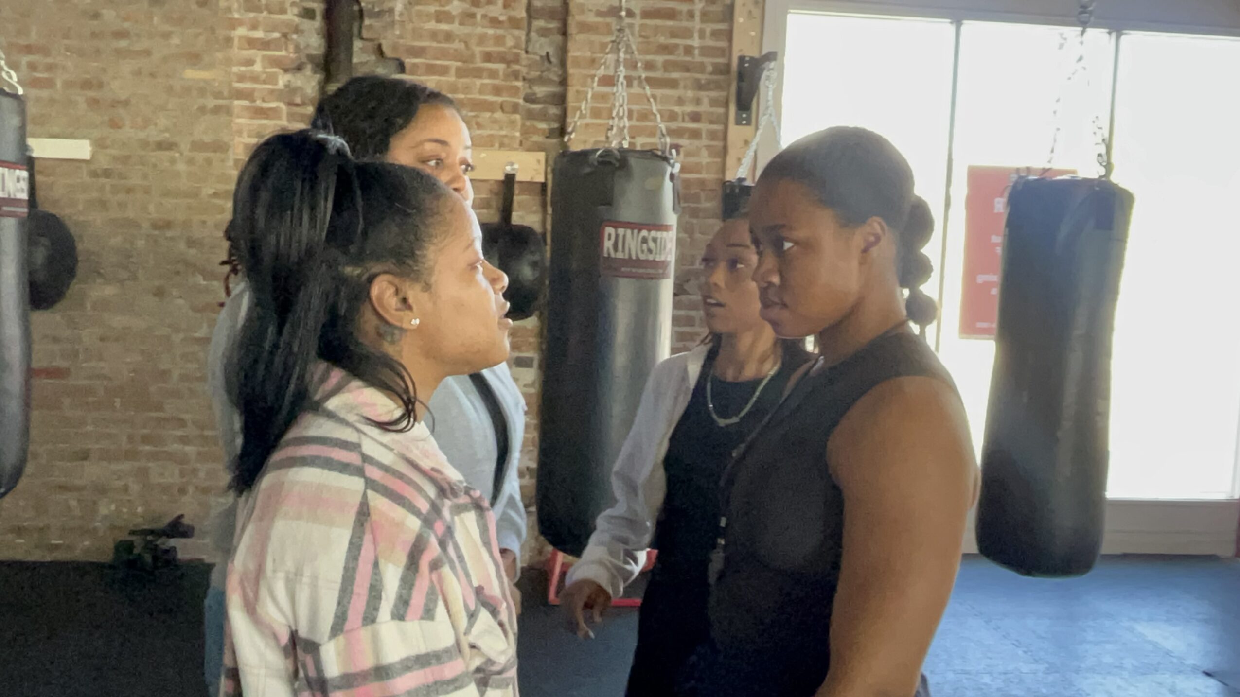 Four women in a gym, standing close and facing each other, with punching bags in the background.