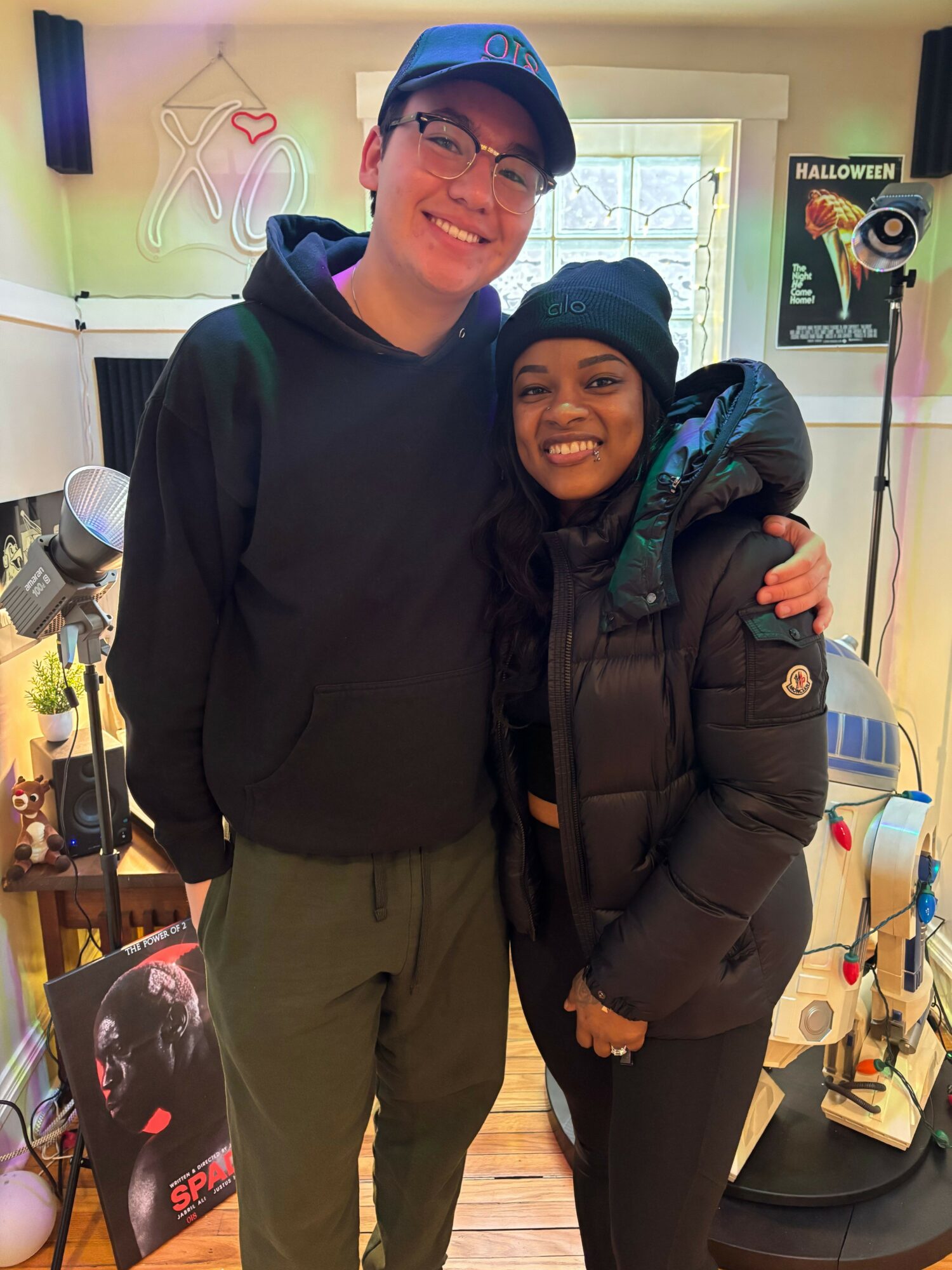 Two people smiling indoors, one wearing glasses and a cap, the other in a black jacket, standing close together.
