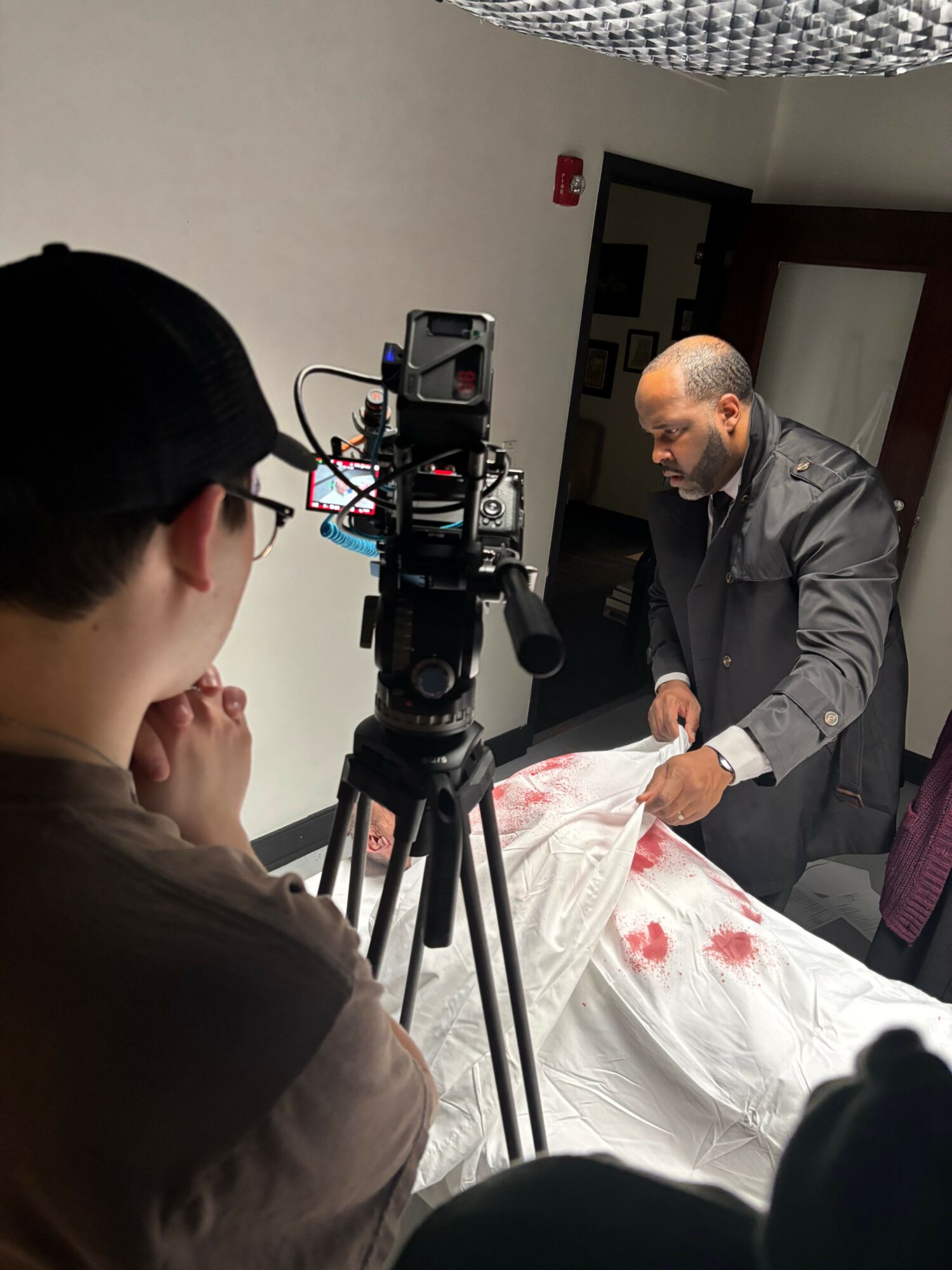 Man in gray suit standing next to a bed with a white sheet, camera setup in foreground, person in brown shirt in foreground.