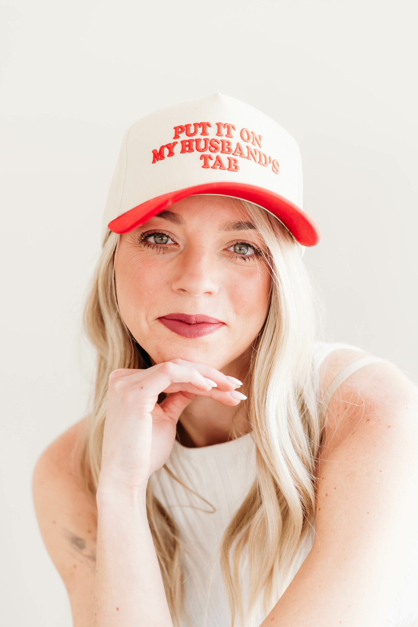Woman with long blonde hair wearing a white and red cap that says 'PUT IT ON MY BUSH AND MY BUBBLY TAB', resting her chin on her hand, smiling.