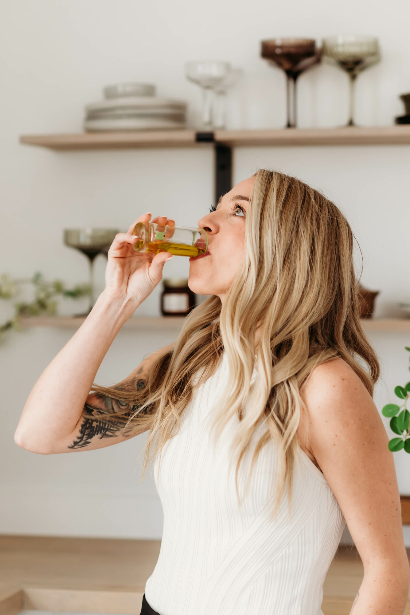 Woman with long blonde hair drinking from a glass bottle in a kitchen, wearing a white sleeveless top.