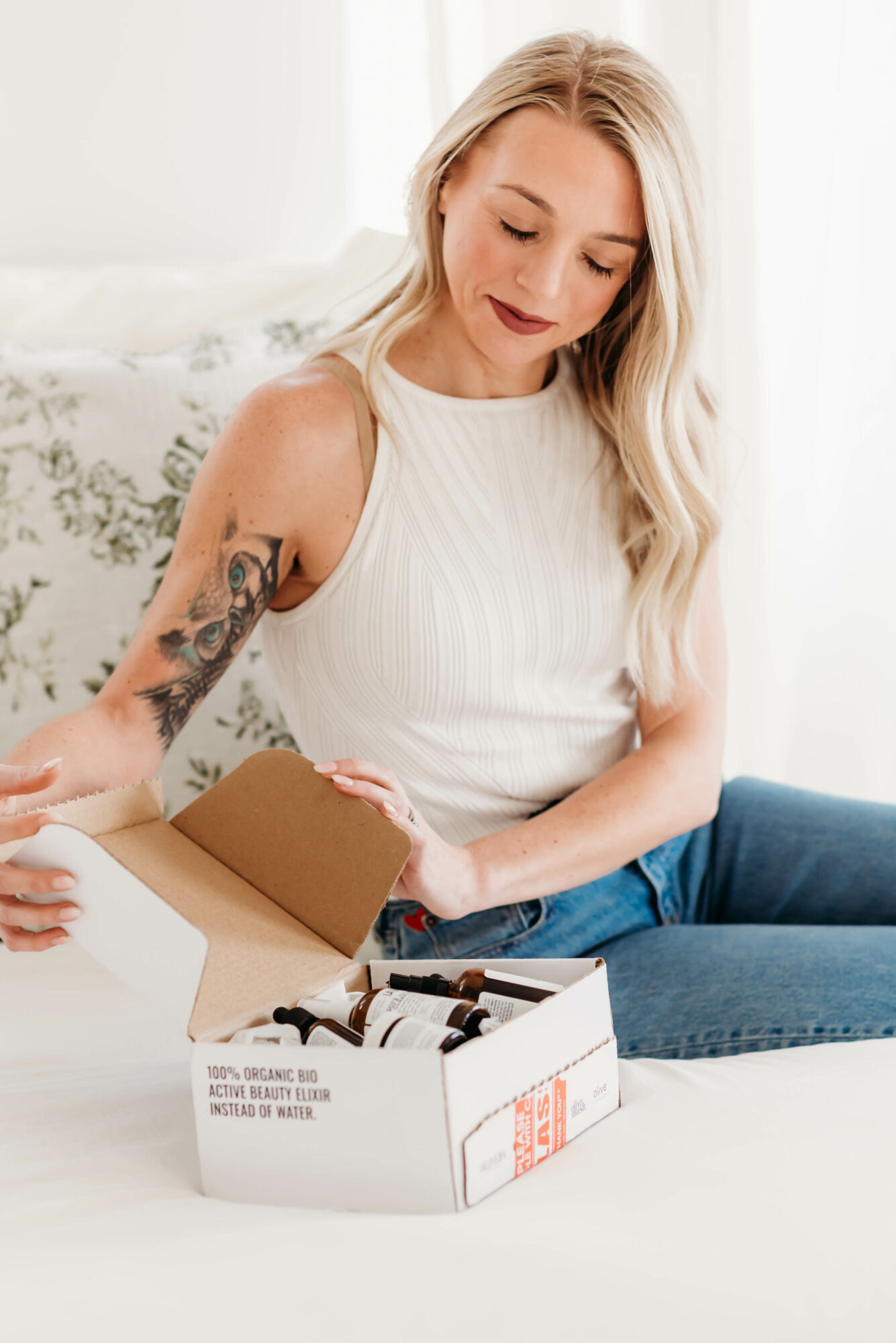 Young woman with long blonde hair and a tattoo on her arm opens a box of chocolates on a white surface.
