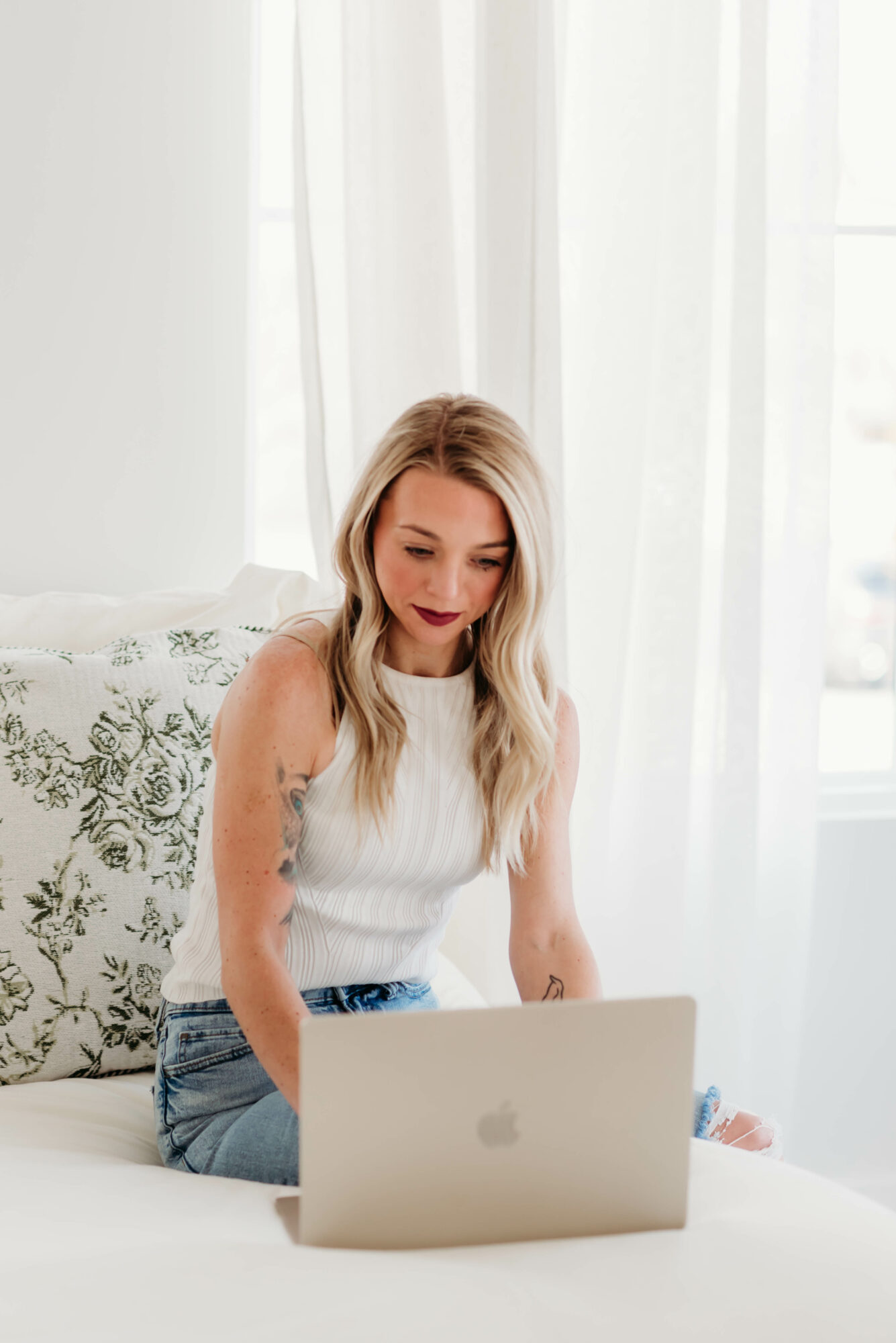 Young woman with long blonde hair using a laptop on a bed in a bright room with white curtains.