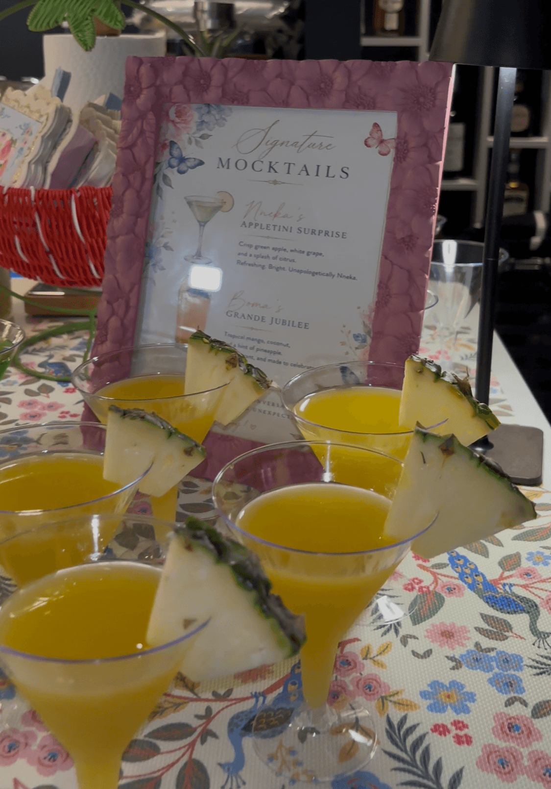 Several yellow cocktails with pineapple garnishes on a floral tablecloth, with a pink menu in the background.
