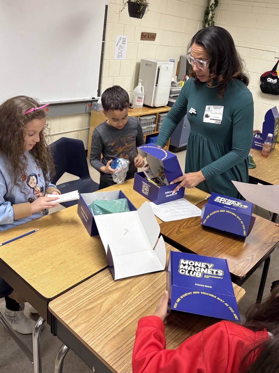 Teacher and children at a table with money-themed activity boxes, some children holding money, in a classroom setting.